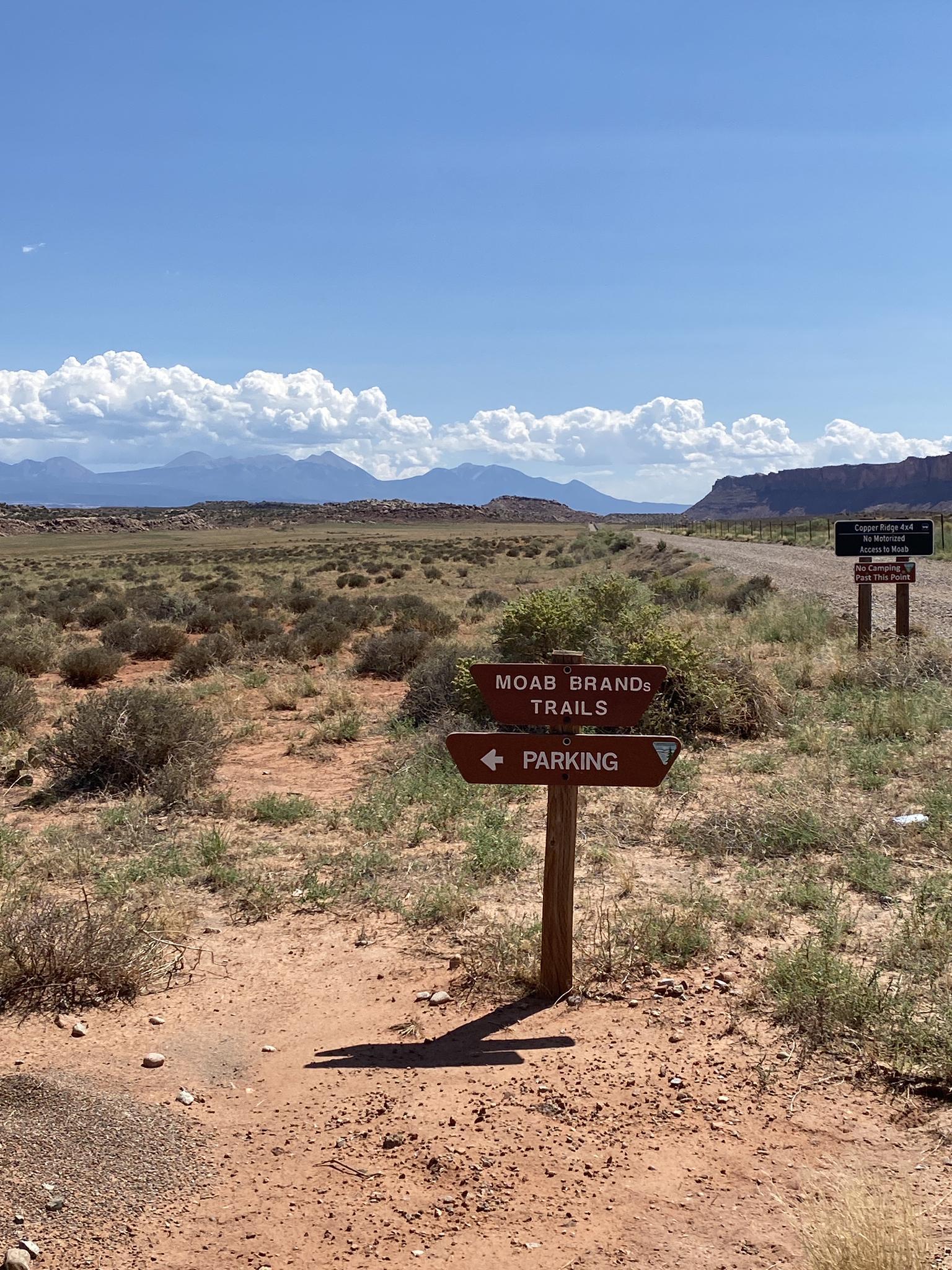 A trail sign in a desert landscape with distant mountains under blue sky.