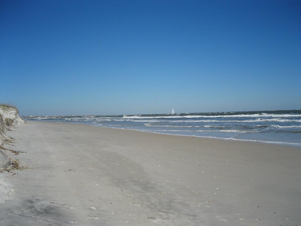 Sunny beach with waves, distant sailboat, and clear blue sky.