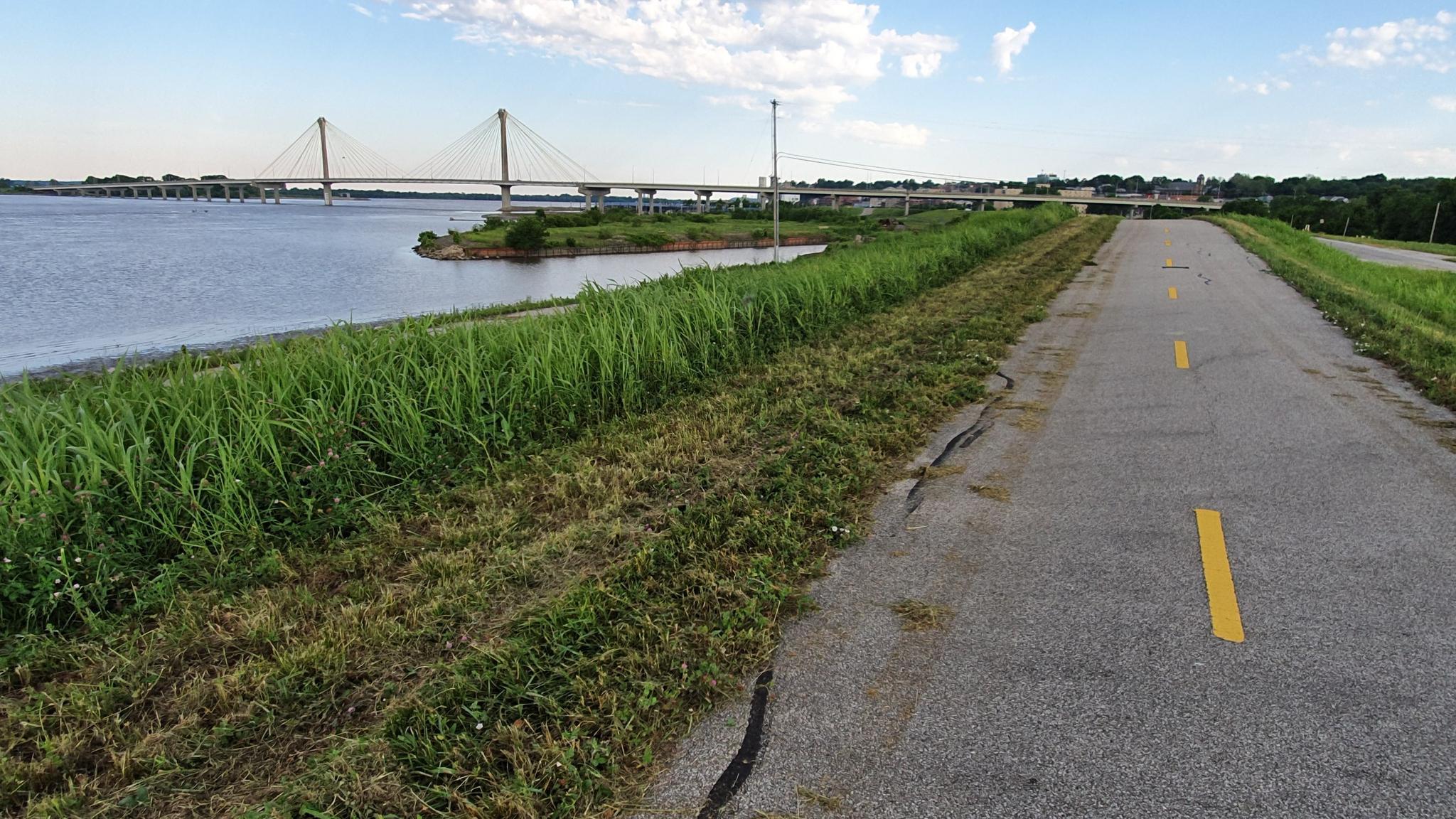 Paved path alongside a wide river with a cable-stayed bridge in daylight.