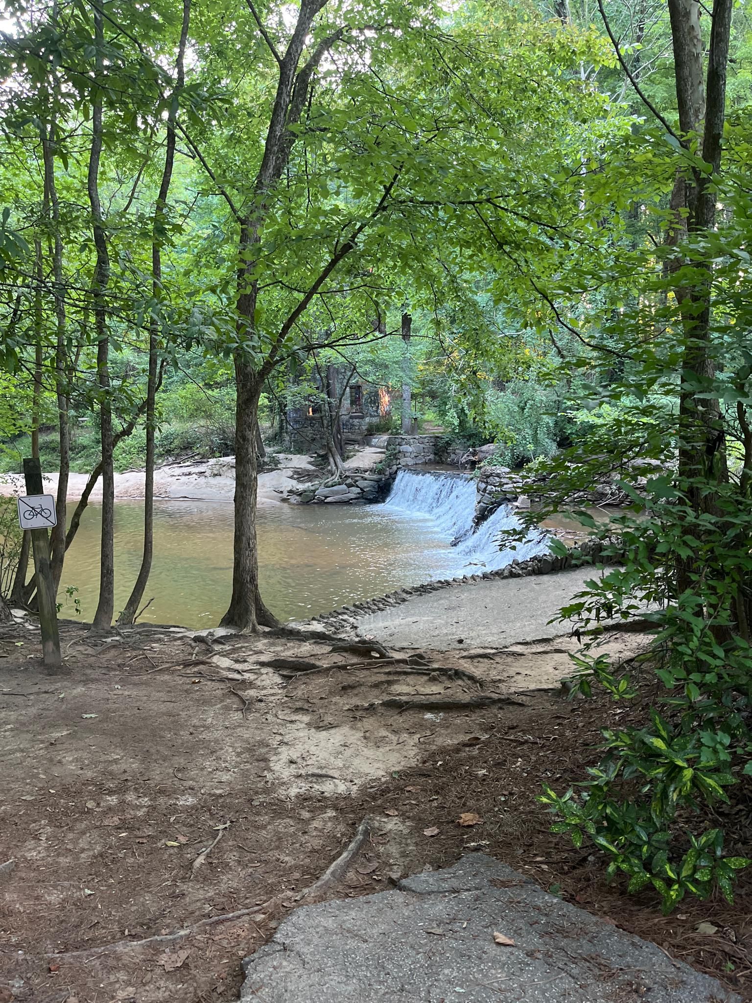A small waterfall cascades into a calm river surrounded by green trees. - Lullwater Loop