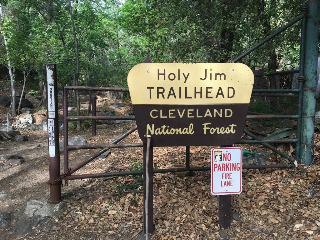 A brown and gold trailhead sign stands before a metal gate in a wooded area.