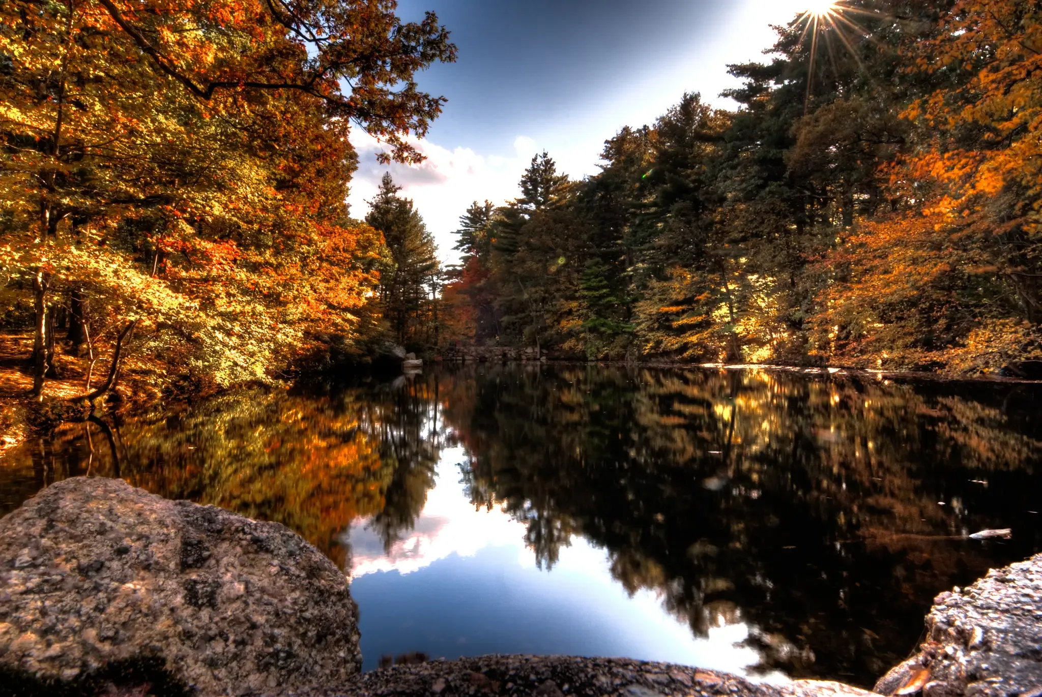 Sunlight streams through colorful autumn trees reflected in a dark lake. - Noanet Woodlands loop