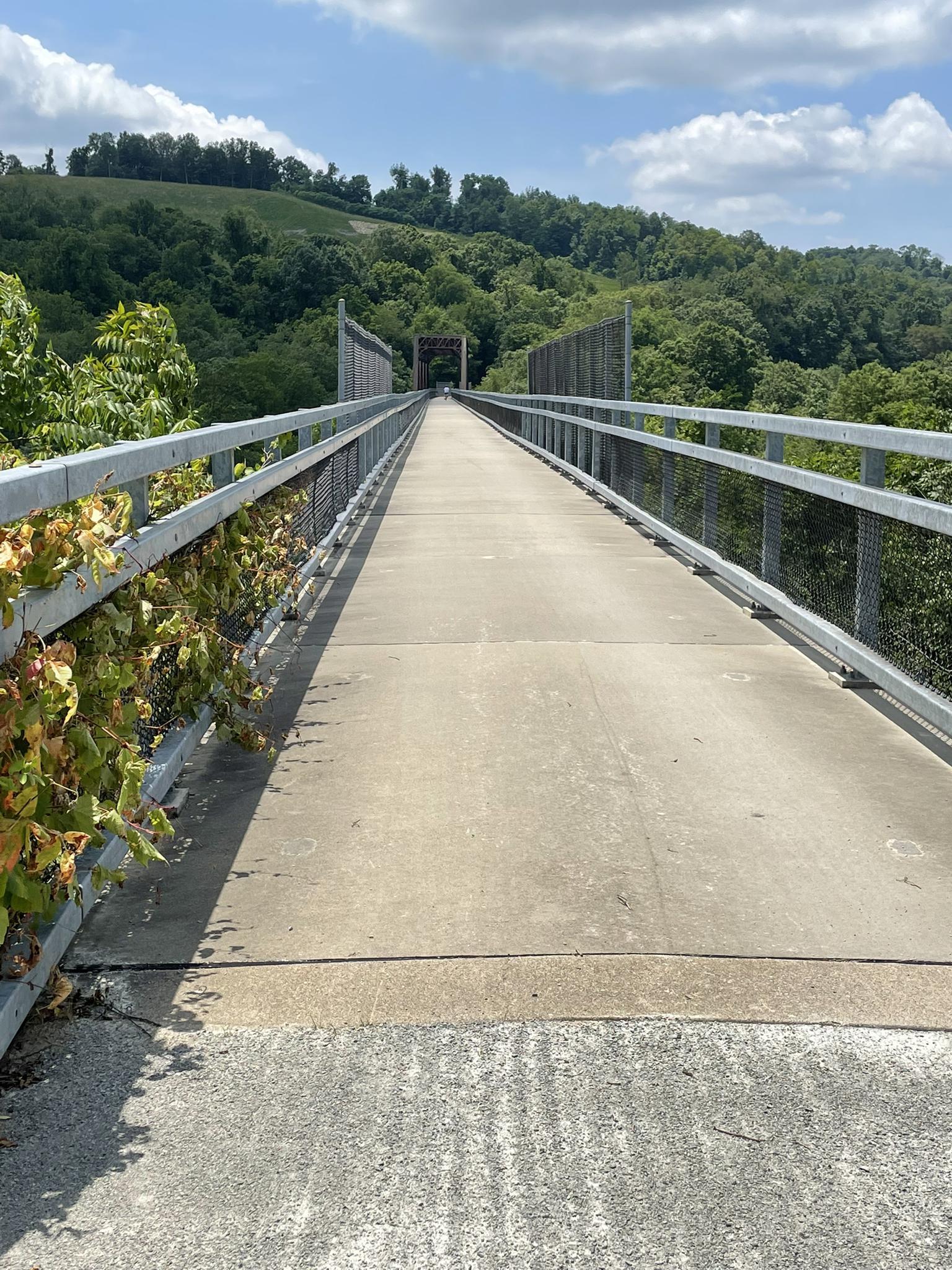 Long concrete path on a bridge with metal railings, leading through green hills on a sunny day.