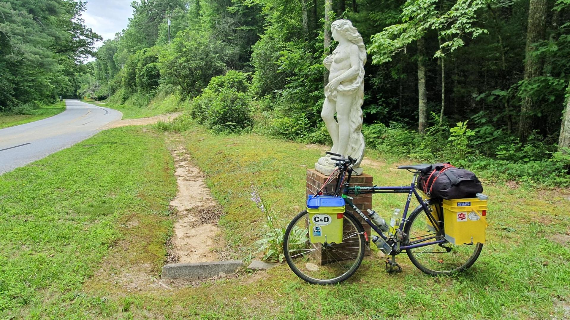 Touring bicycle with panniers parked beside a white classical statue on a grassy roadside.