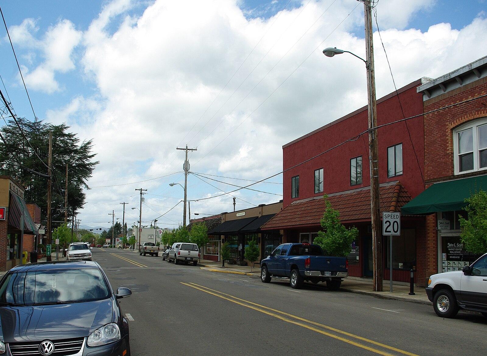 A quiet town street with buildings, parked cars, and a cloudy sky.