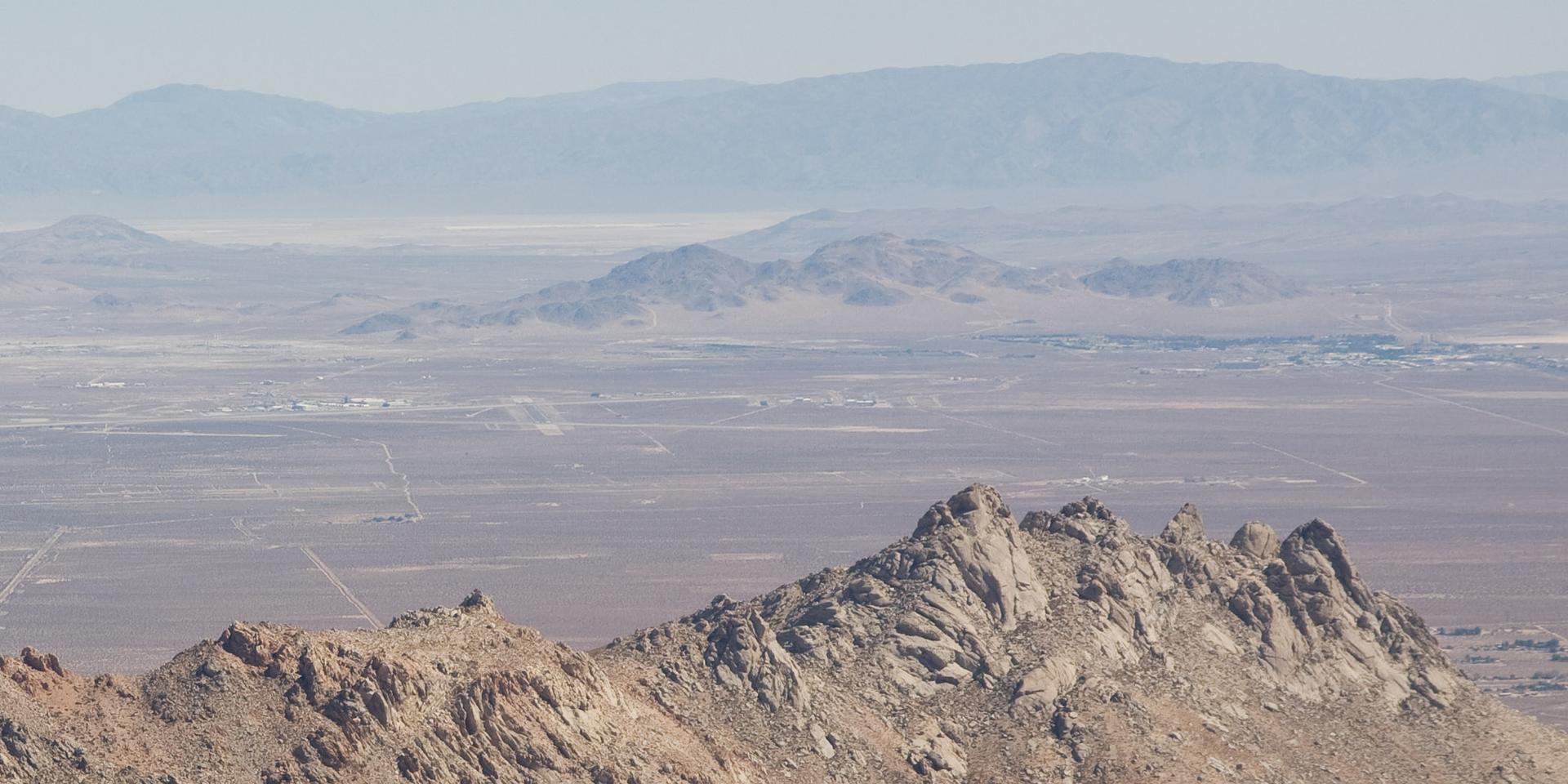 Rocky peaks overlook a vast, hazy desert valley under bright skies.