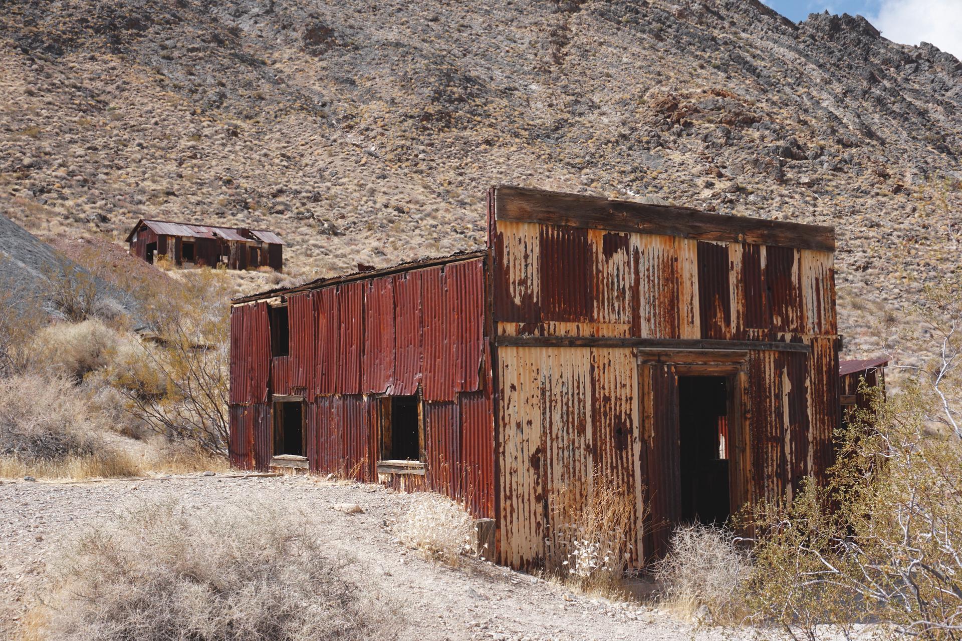 Two rusty, dilapidated corrugated metal buildings on a dry, rocky hillside.