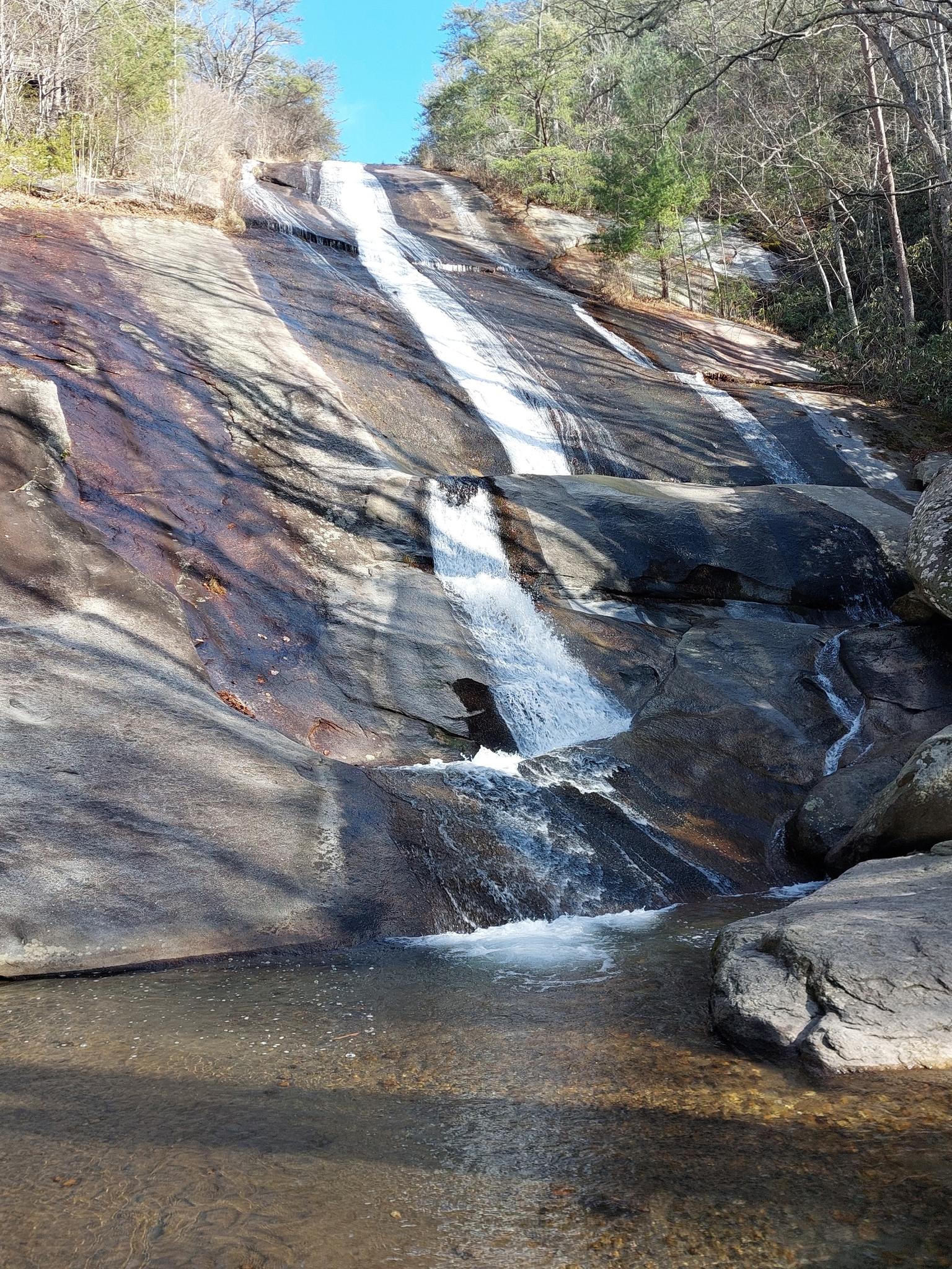 A wide waterfall cascades down a large, smooth rock face into a pool.