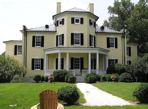 Large yellow house with a white columned porch, green lawn, and bright sunlight.