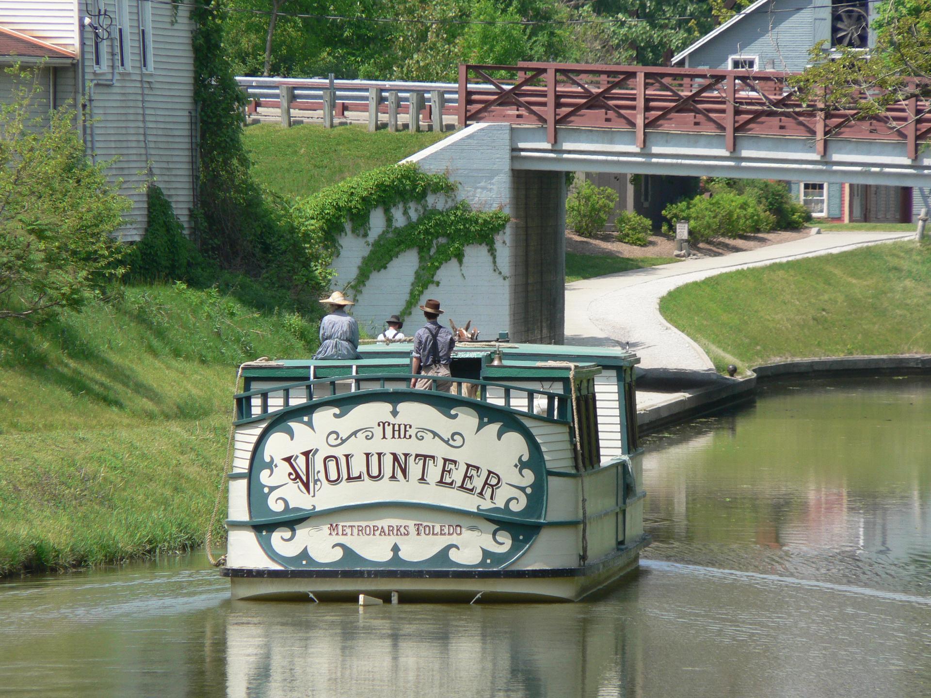 People ride a historic canal boat named "The Volunteer" under a bridge.