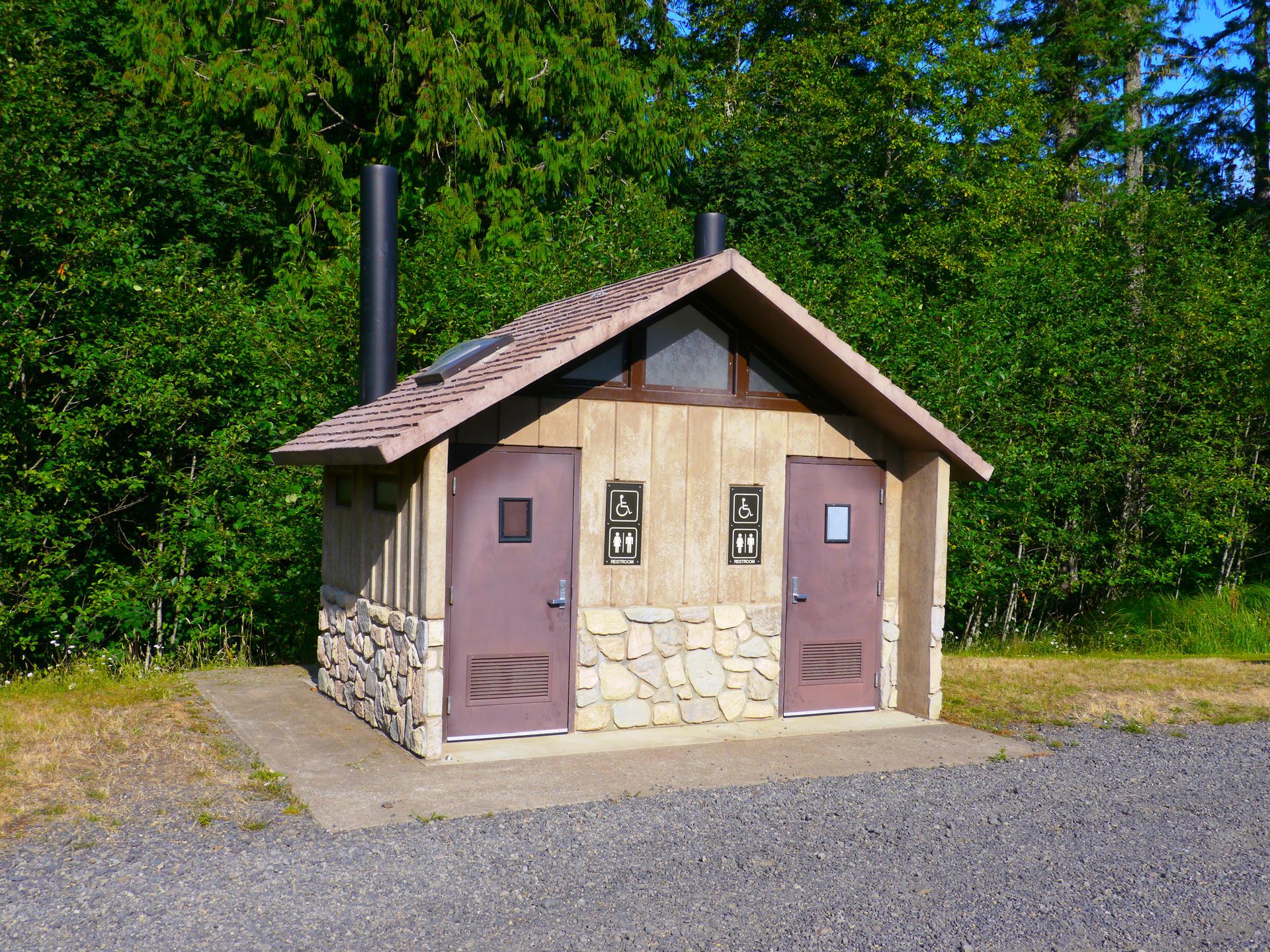 A rustic restroom building with two doors, surrounded by green trees on a sunny day.