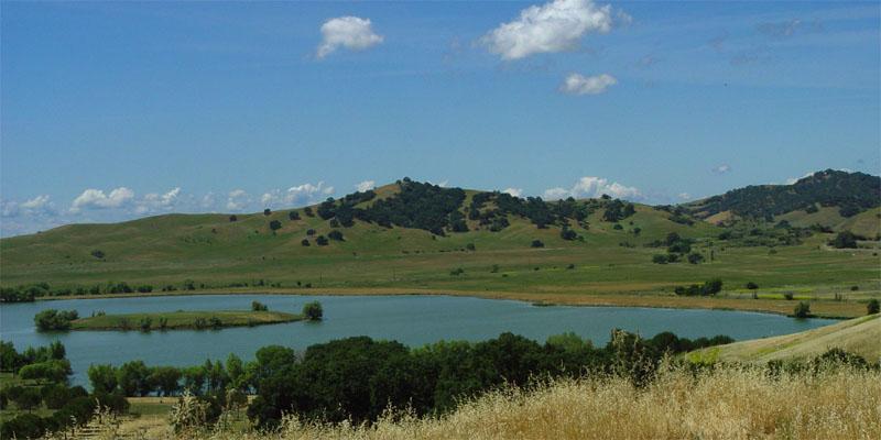 A serene lake surrounded by rolling green hills under a bright blue sky.