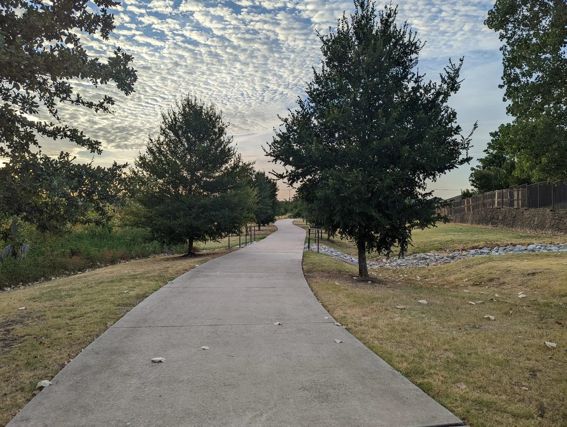 A paved path winds through grassy areas with trees under a cloudy sky.