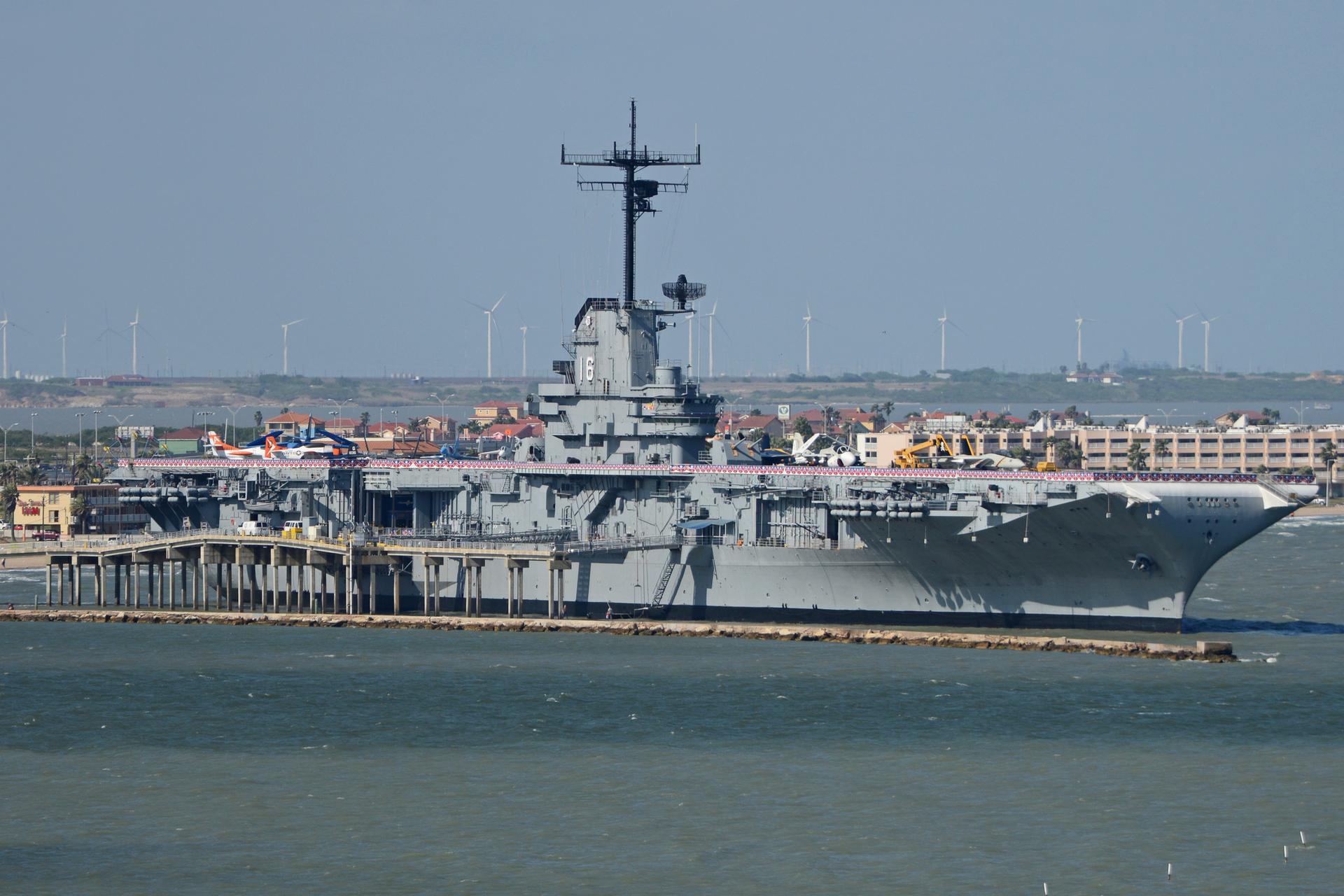 A large gray aircraft carrier is docked by a city with wind turbines.