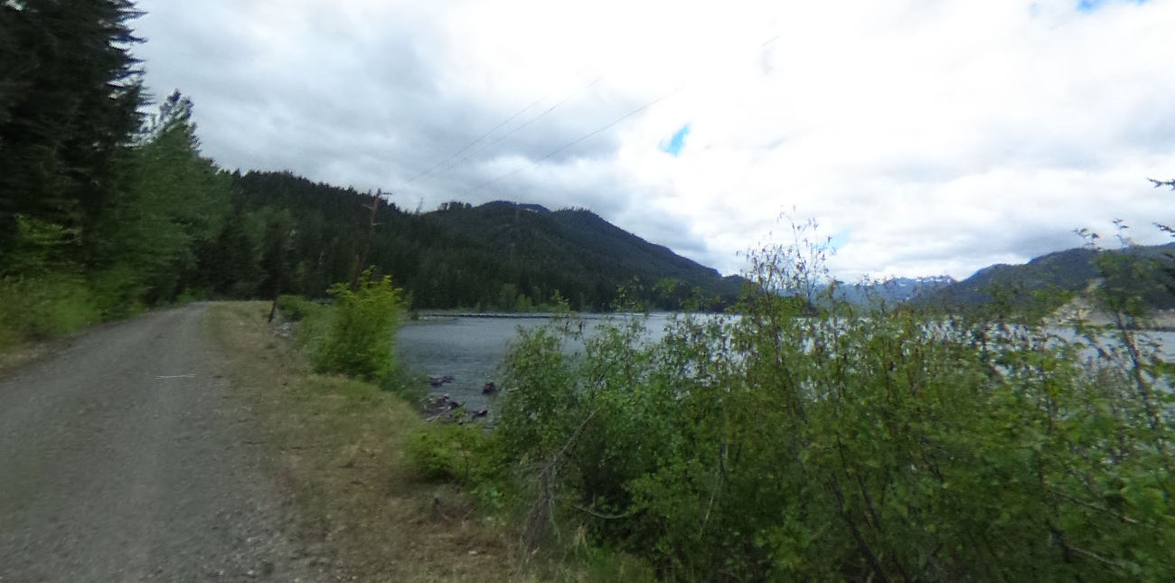 Gravel road next to a lake, with dense forest and mountains under cloudy sky.