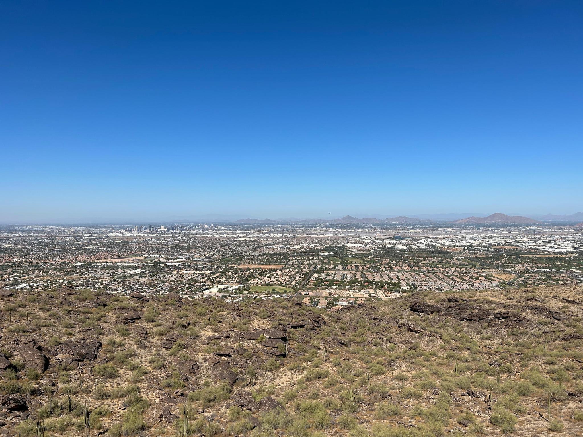 Expansive city view from a rocky desert hill under a clear blue sky.