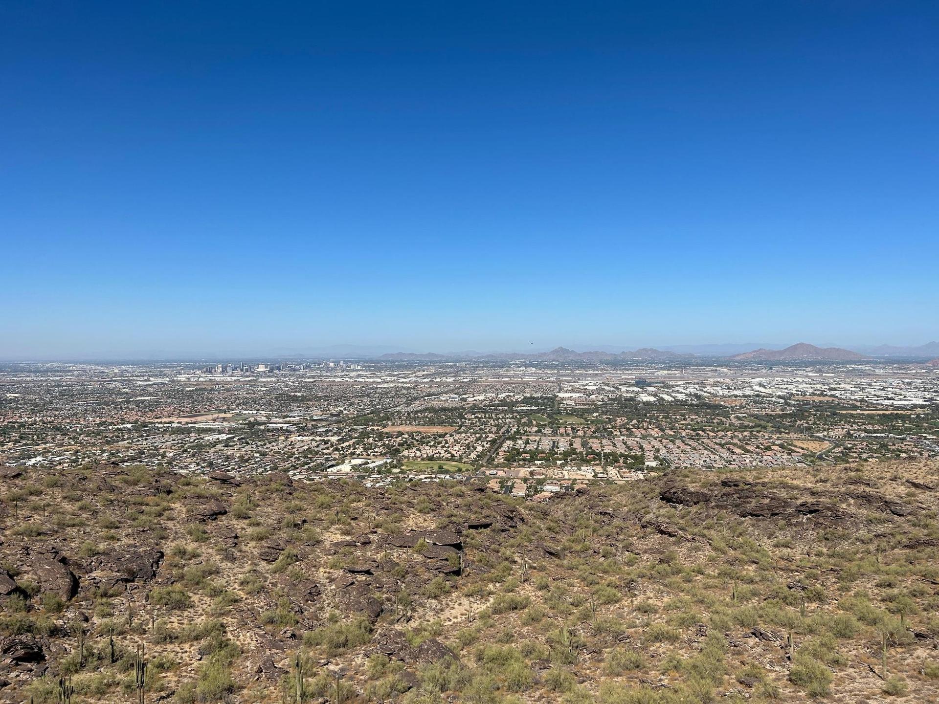 Expansive city view from a rocky desert hill under a clear blue sky.