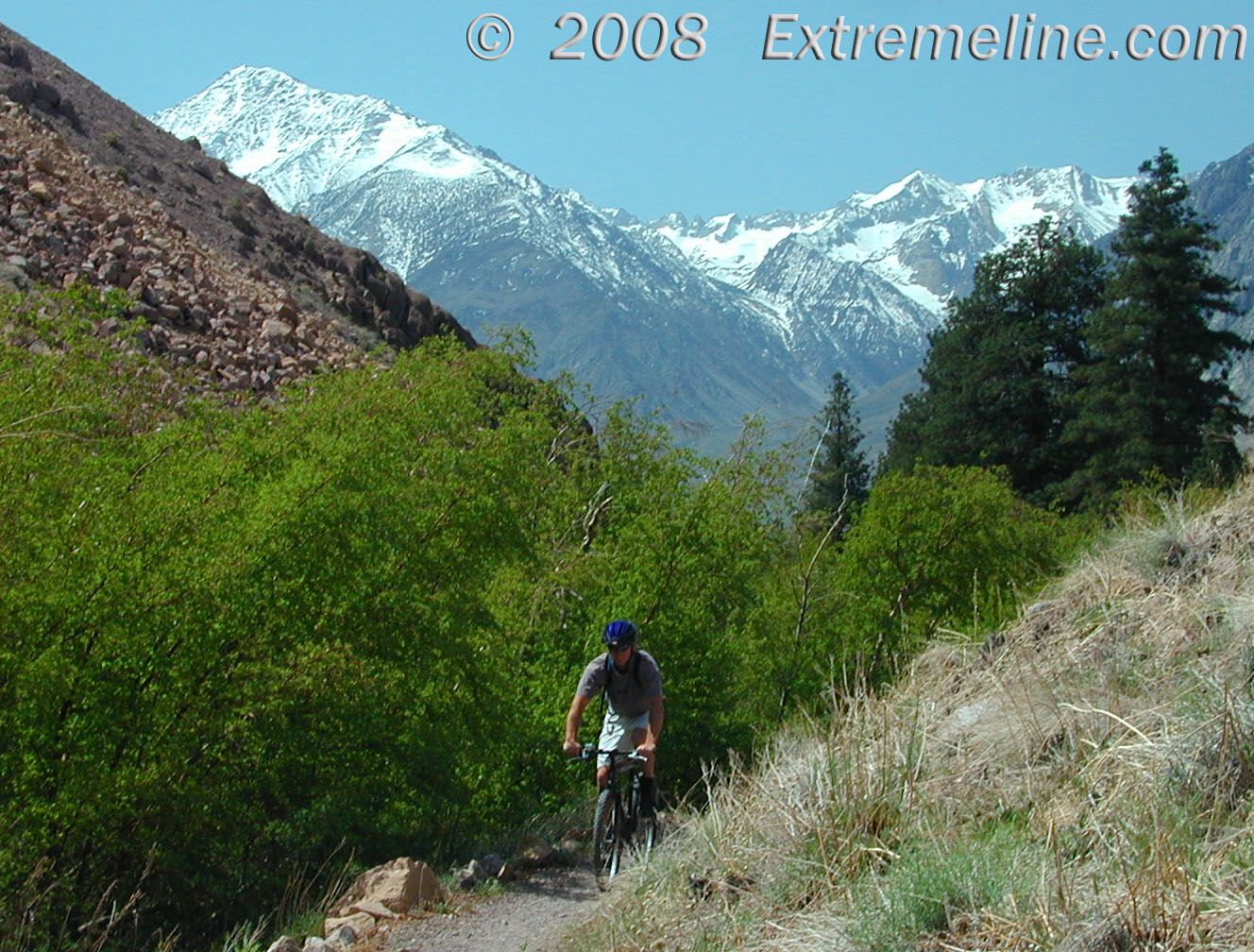 Mountain biker on a dirt trail with snow-capped mountains in bright sunlight. - Lower Rock Creek Trail
