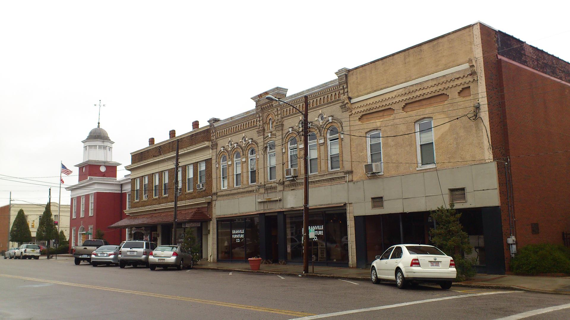 Historic brick buildings and a red cupola building line a street with parked cars.