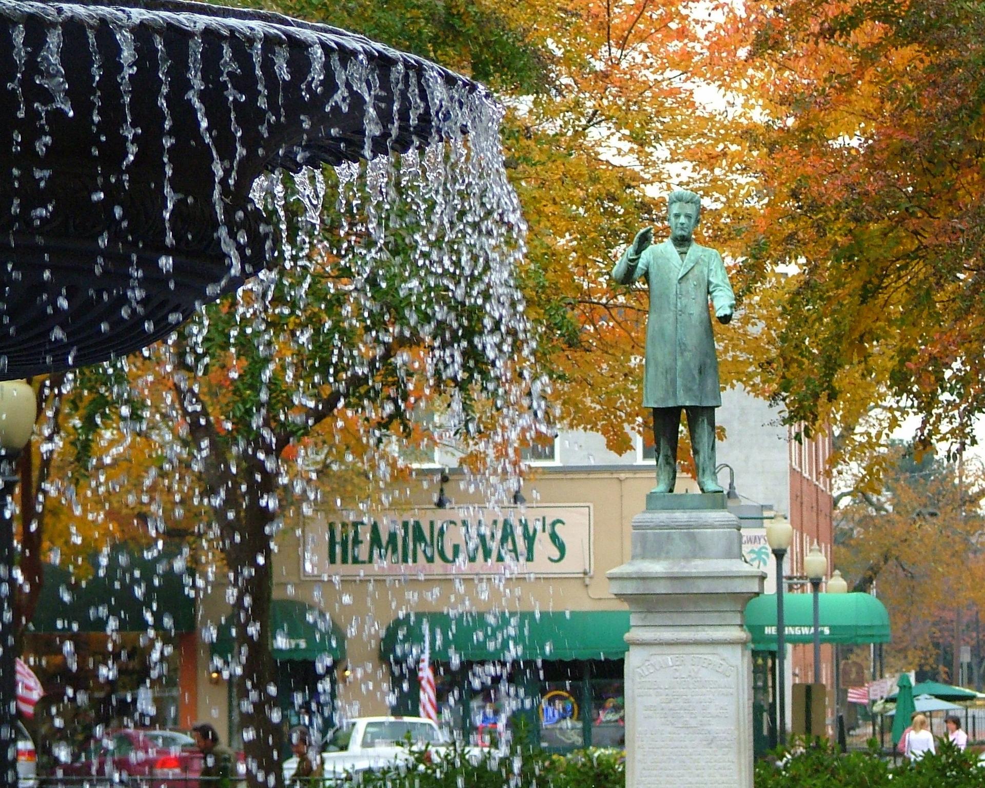 Water cascades from a fountain near a bronze statue and autumn trees.