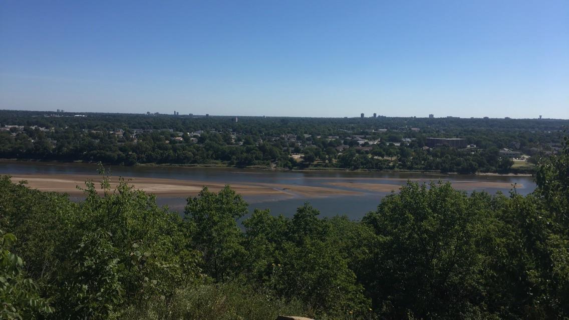 Wide river with sandbars, green trees, and a distant city under clear blue sky.