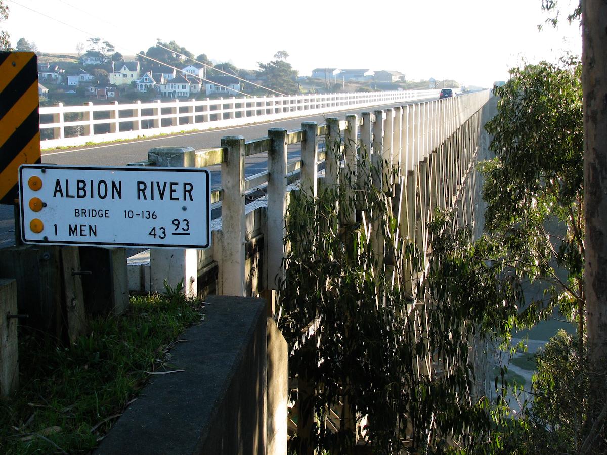 Long bridge over water with a sign, leading to a distant town, bright sunlight.