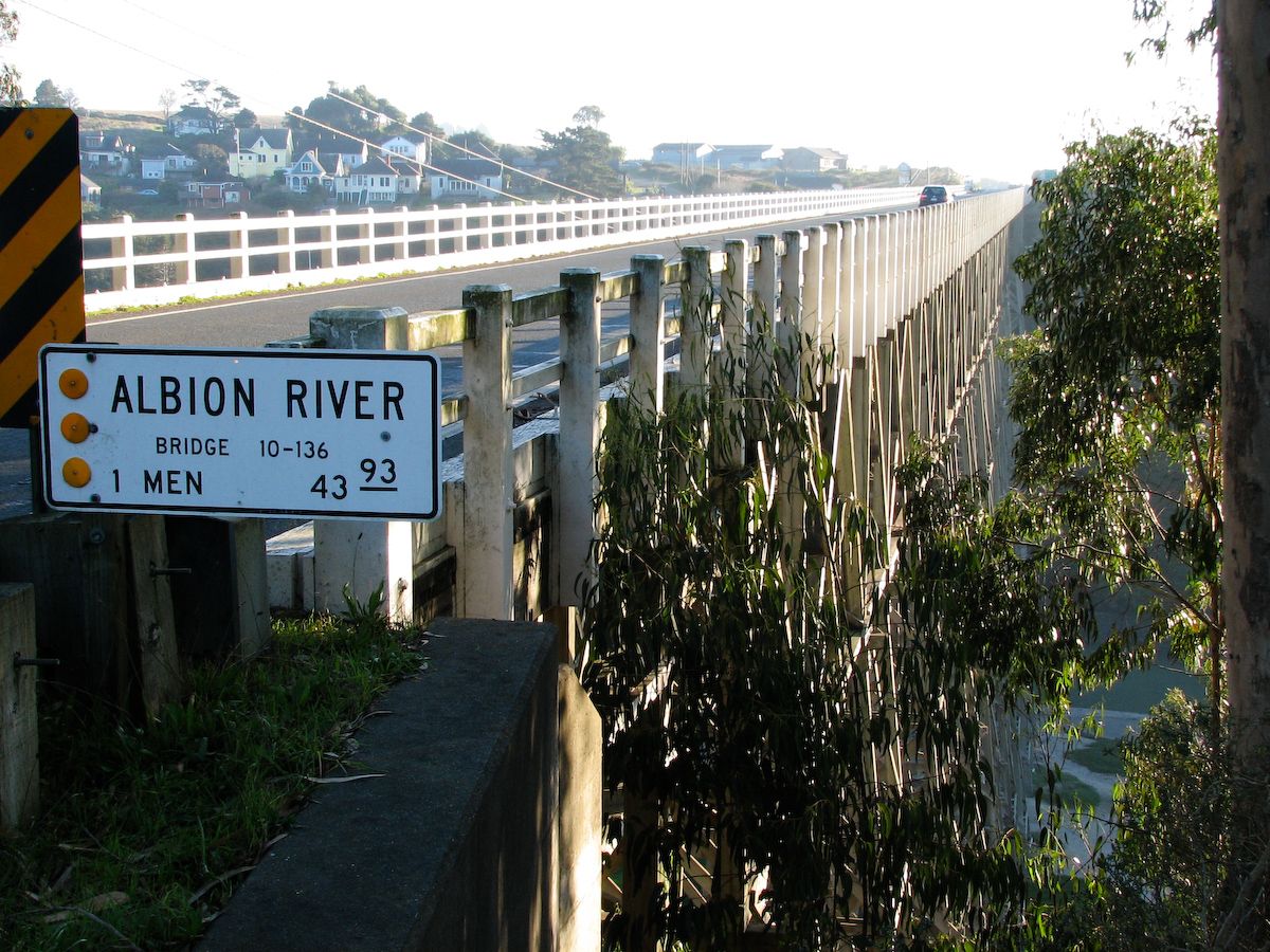 Long bridge over water with a sign, leading to a distant town, bright sunlight. - Mendocino - Comptche loop