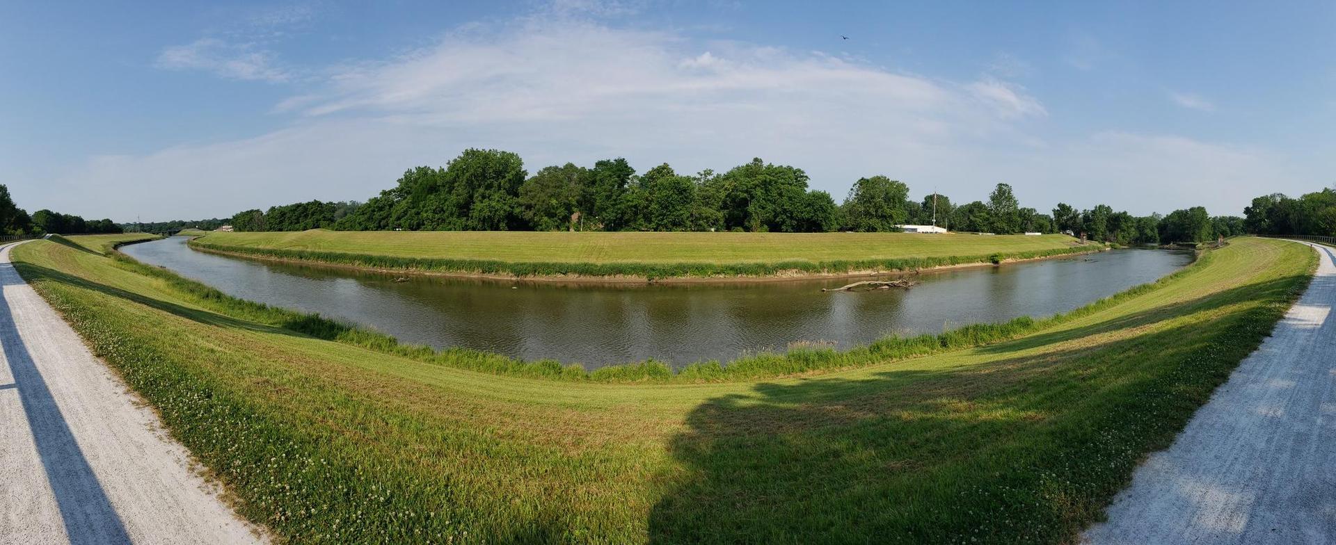 Wide river with green grassy banks and distant trees under a blue sky.