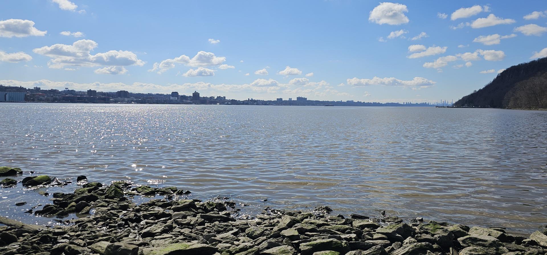 Sunny river view from a rocky shore with a distant city skyline.