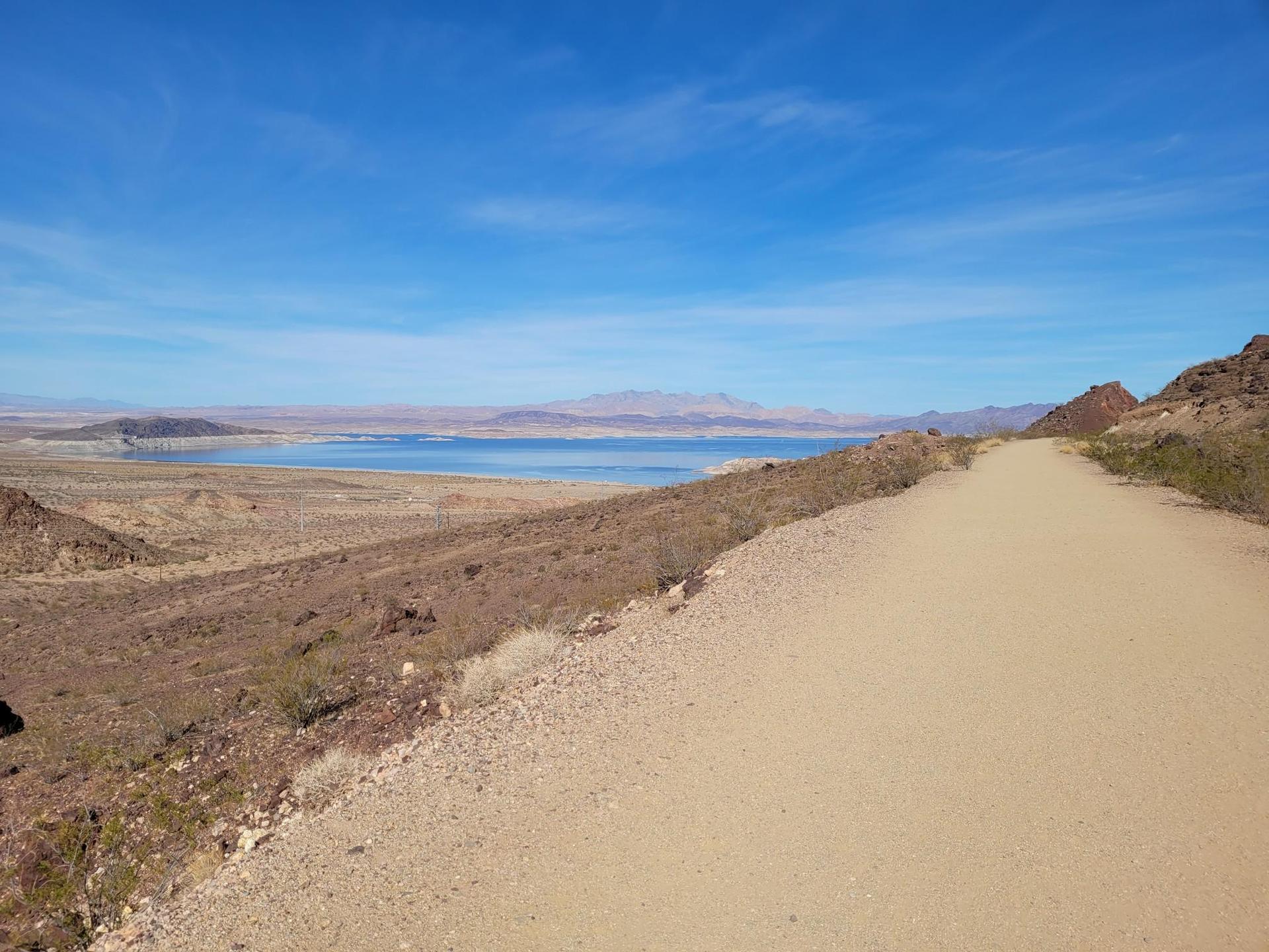 Wide dirt path overlooking a vast desert landscape with a distant lake.