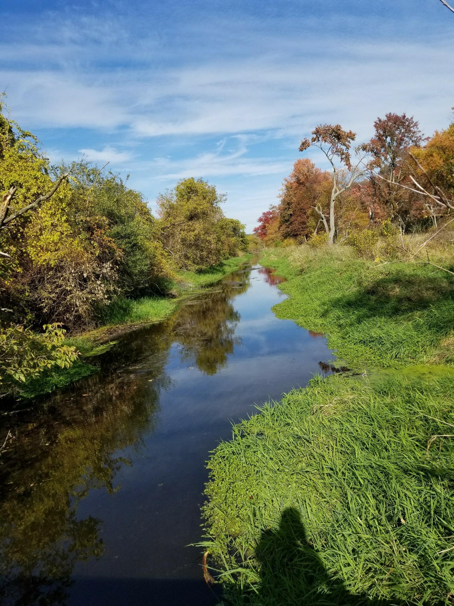A narrow river with green banks and autumn trees under a blue sky.