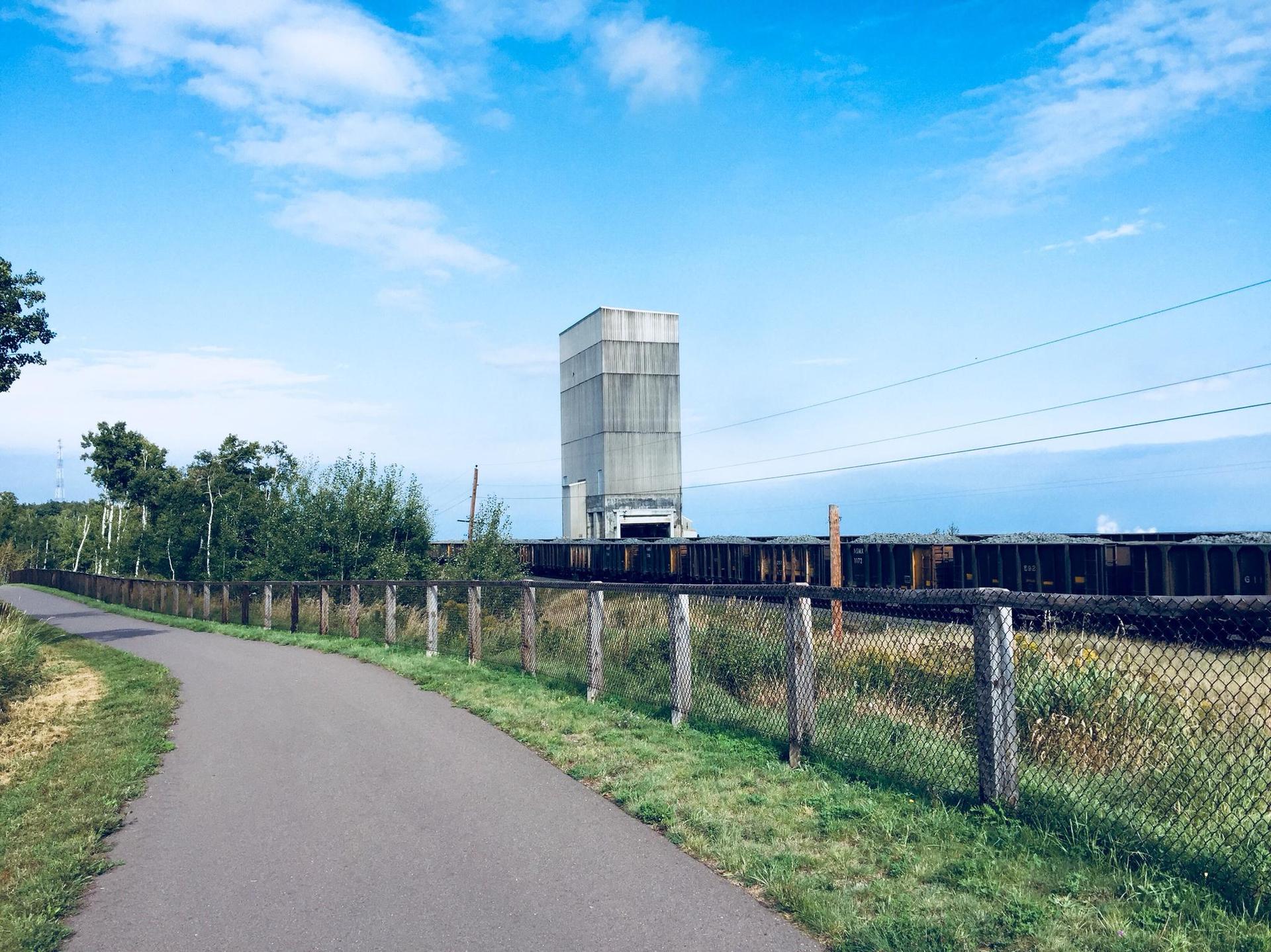 Paved path, fence, industrial building, and train cars under a bright blue sky.