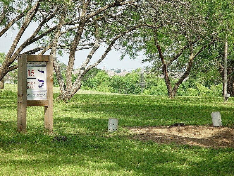 Wooden disc golf sign and basket on a bright, green, tree-filled course. - Sharon Prete Main Street Plaza – Old Settlers Park loop from Round Rock