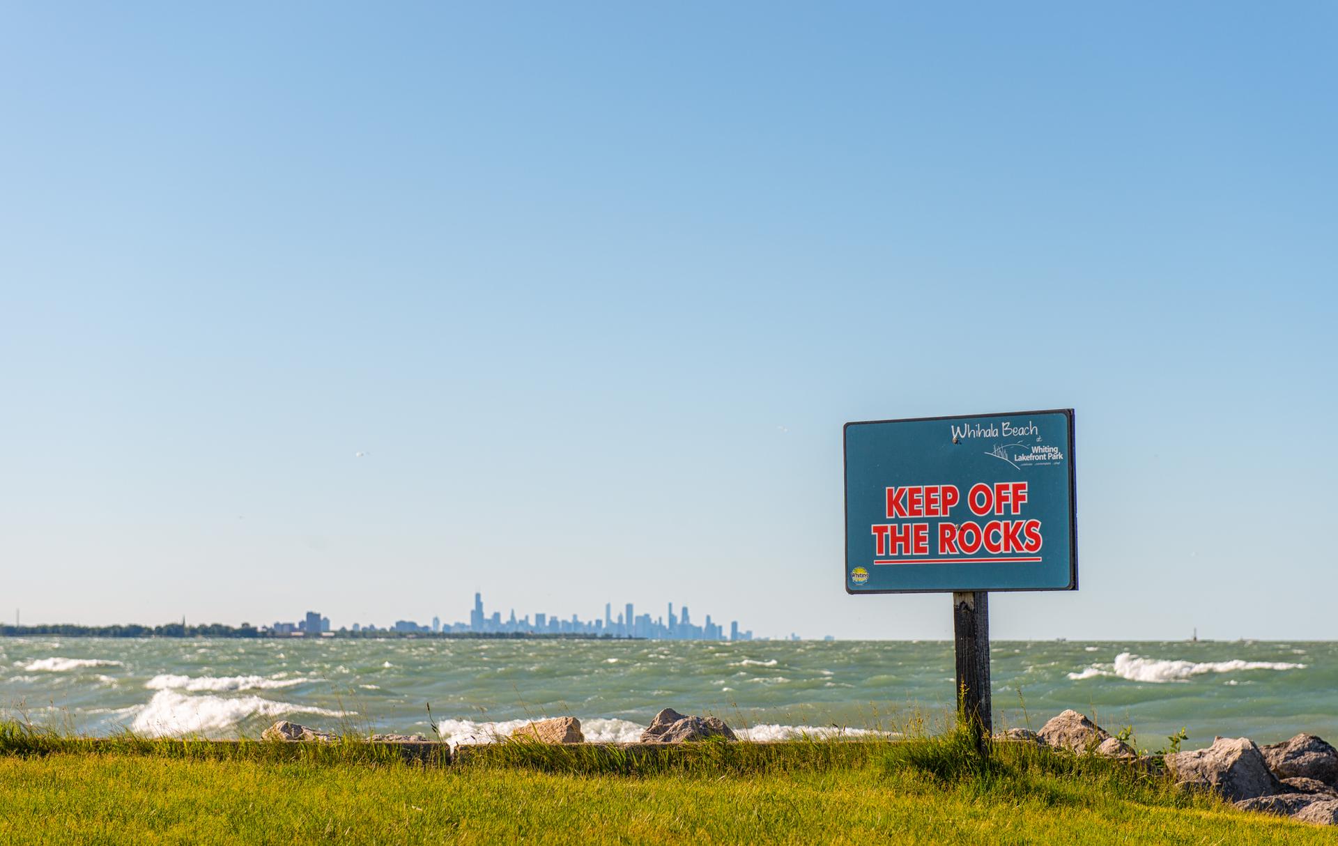 A "KEEP OFF THE ROCKS" sign by choppy water with a distant city skyline.