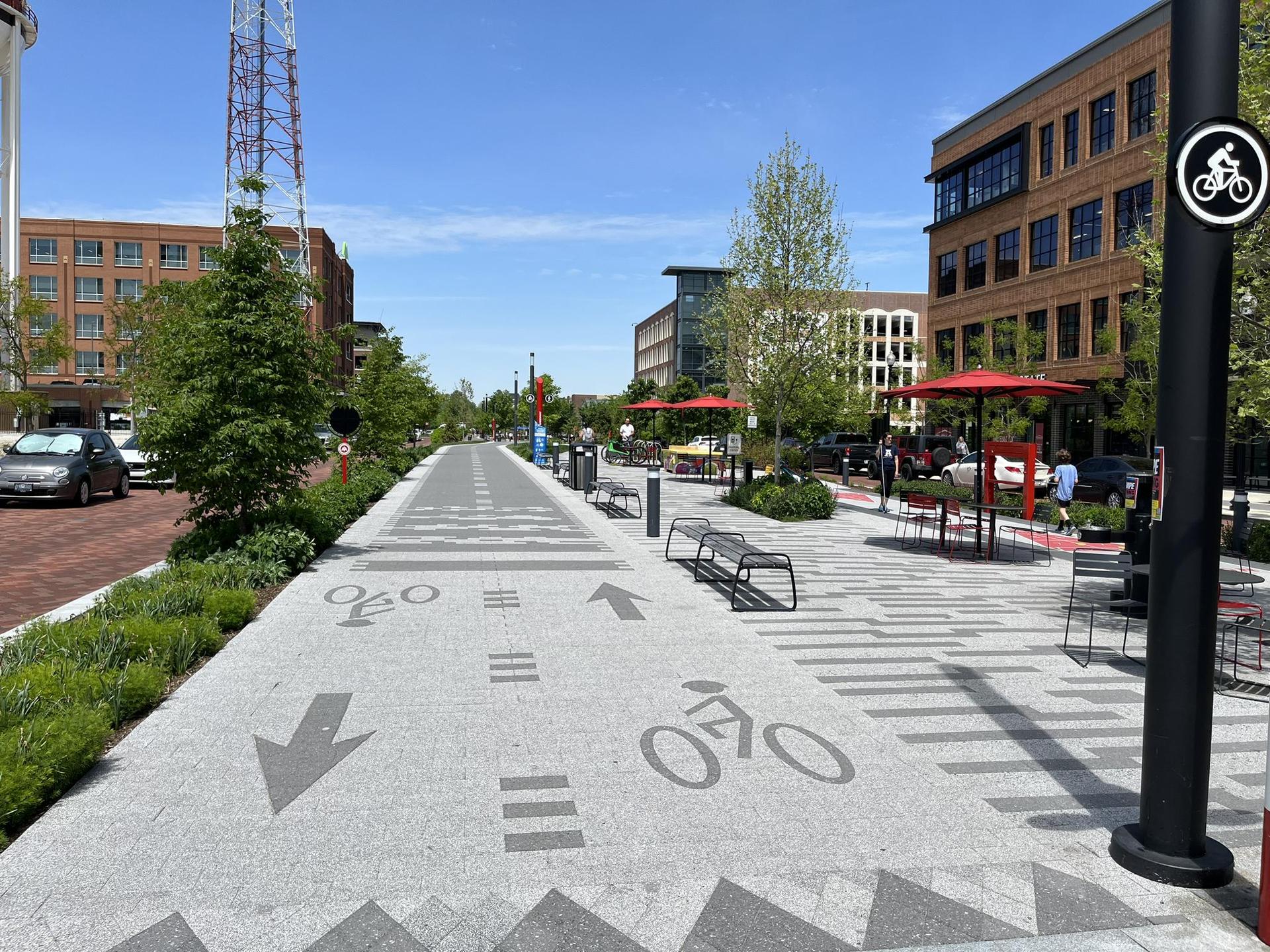 Wide urban pathway with bike lane markings, modern buildings, and outdoor seating.