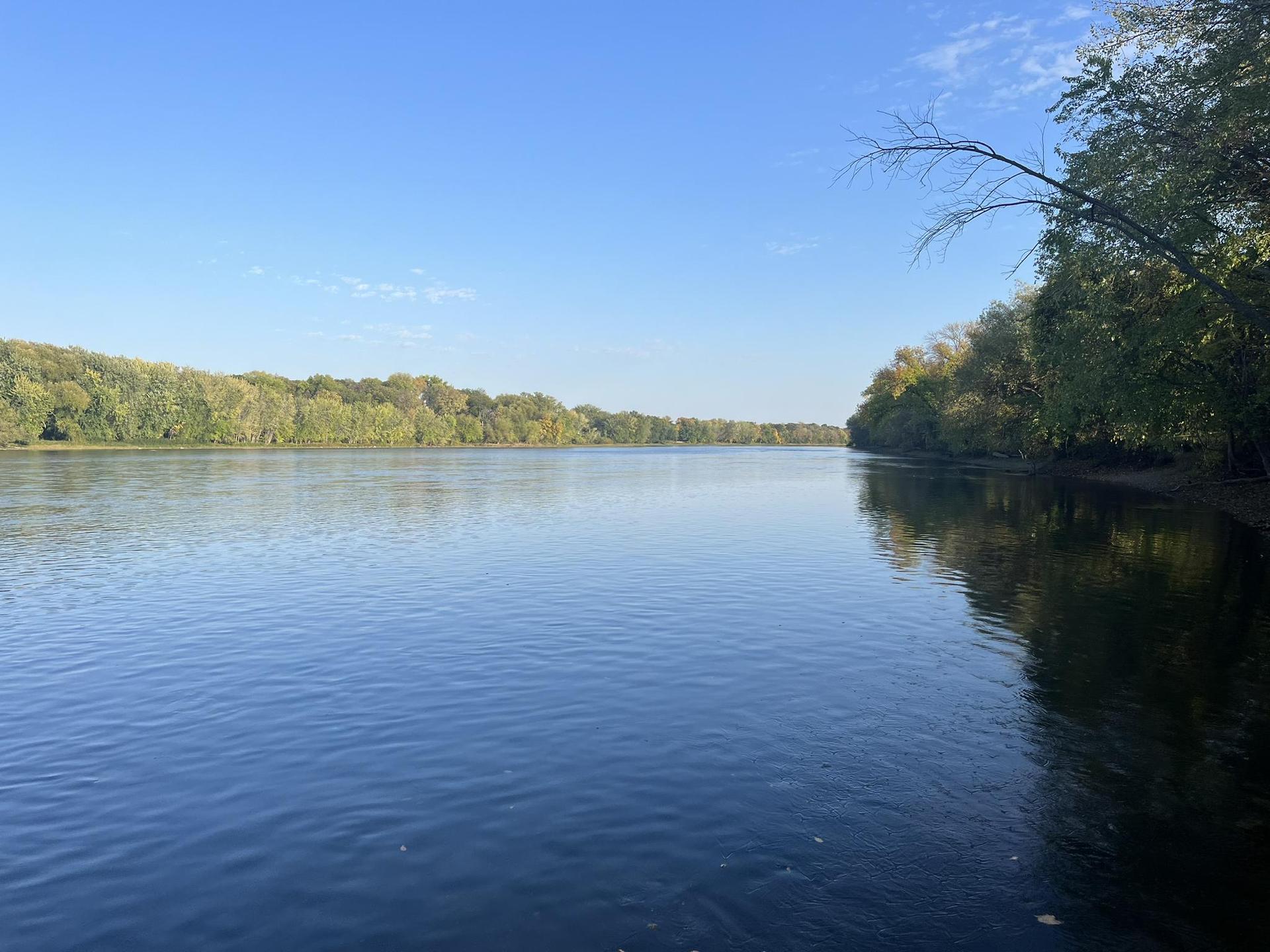 A wide, calm river flows between lush green tree-lined banks under a clear blue sky.