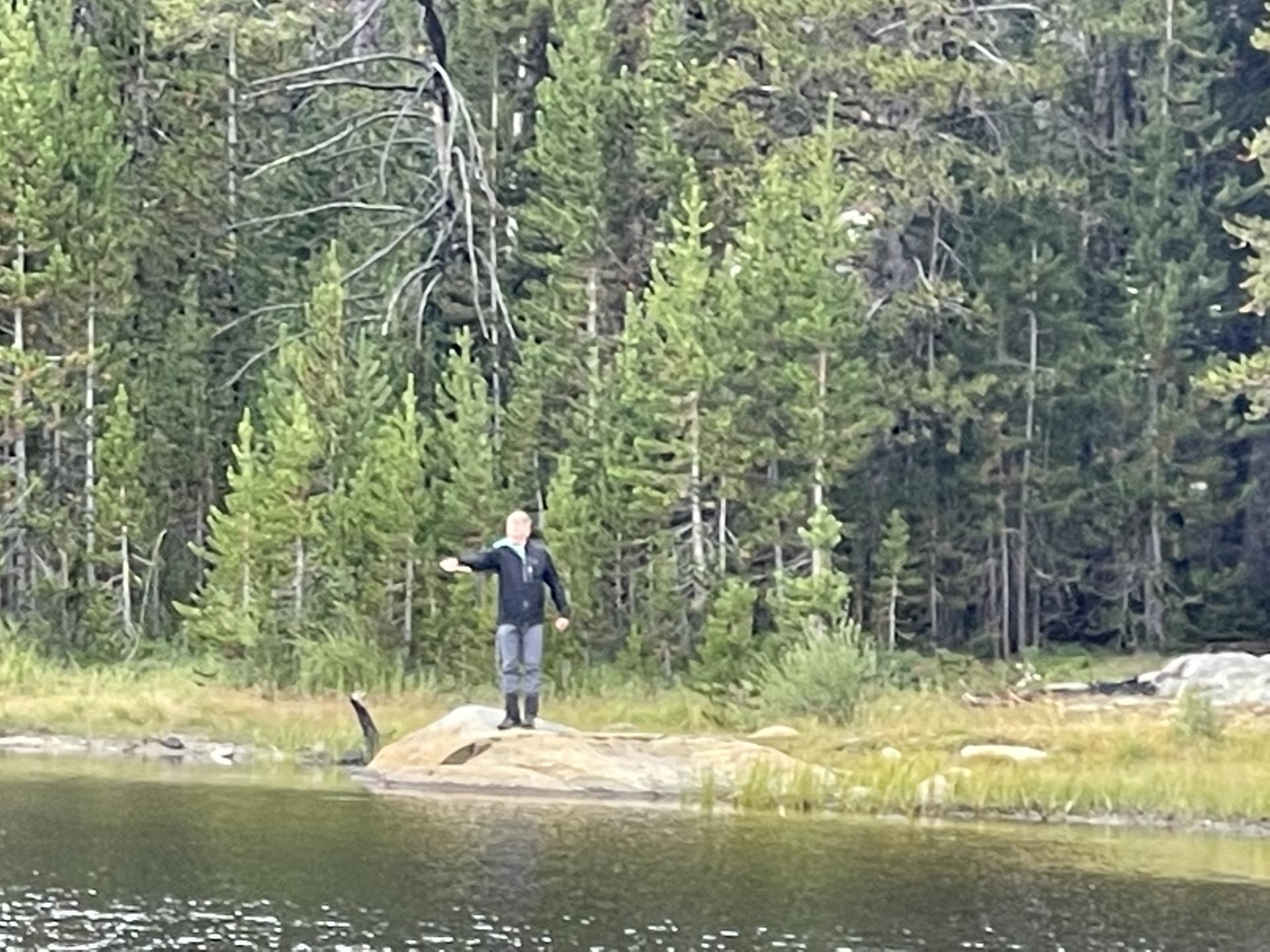 Person on a rock in a lake, arm outstretched, with a dense pine forest.