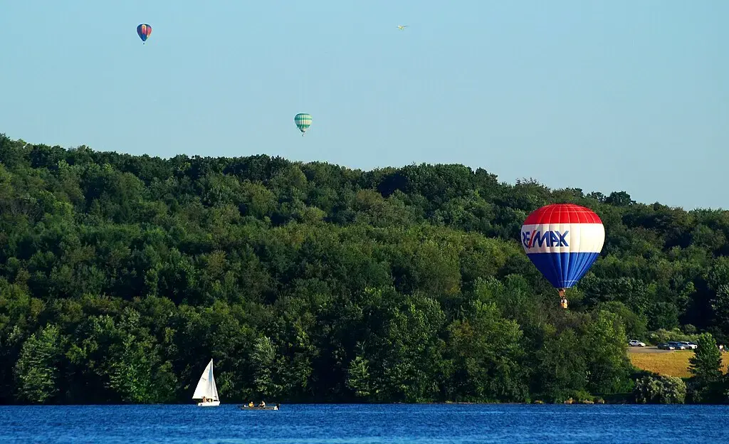 Hot air balloons and a sailboat on a lake with forested hills.