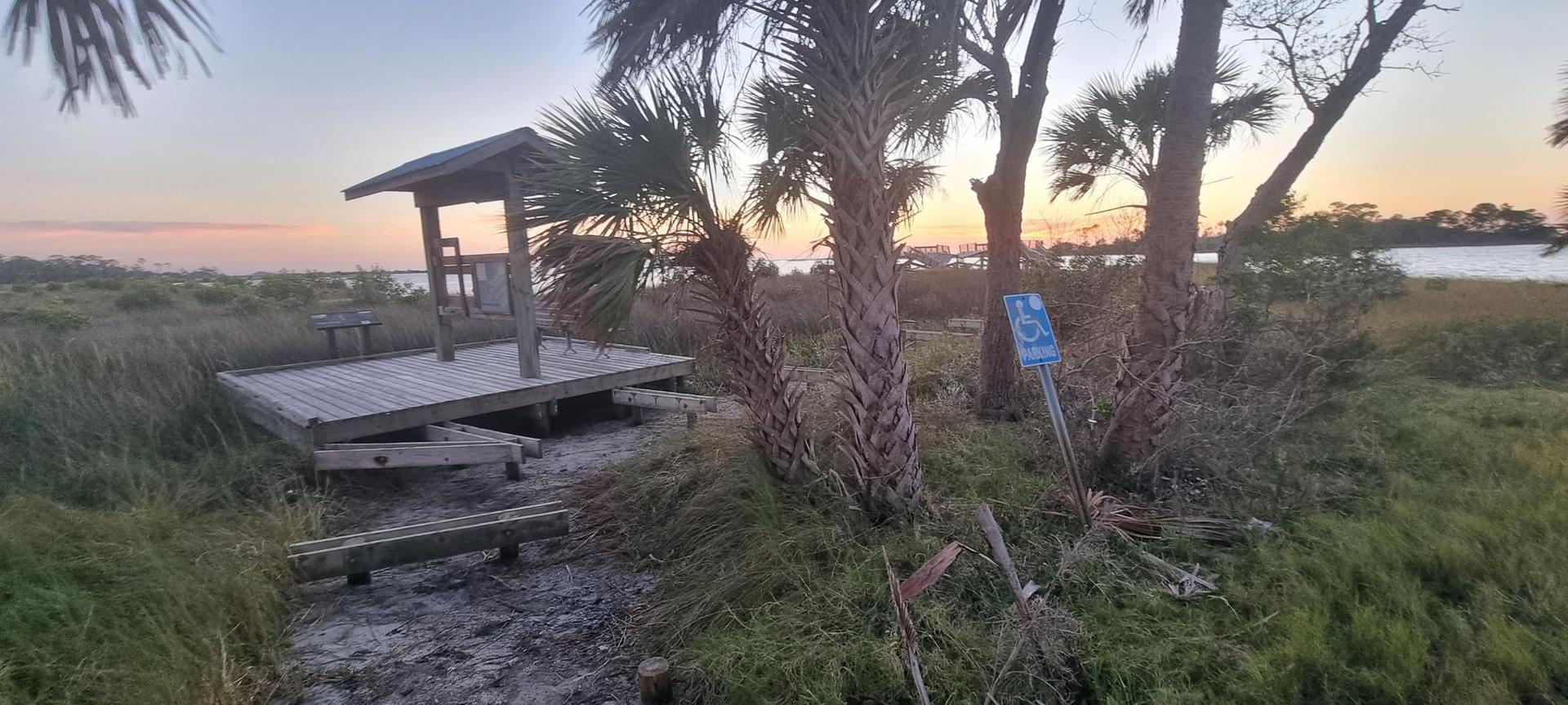 Wooden platform and shelter with palm trees at sunset by a body of water.