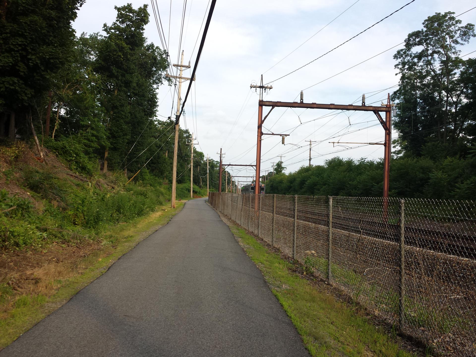 Paved path alongside train tracks and power lines under an overcast sky.