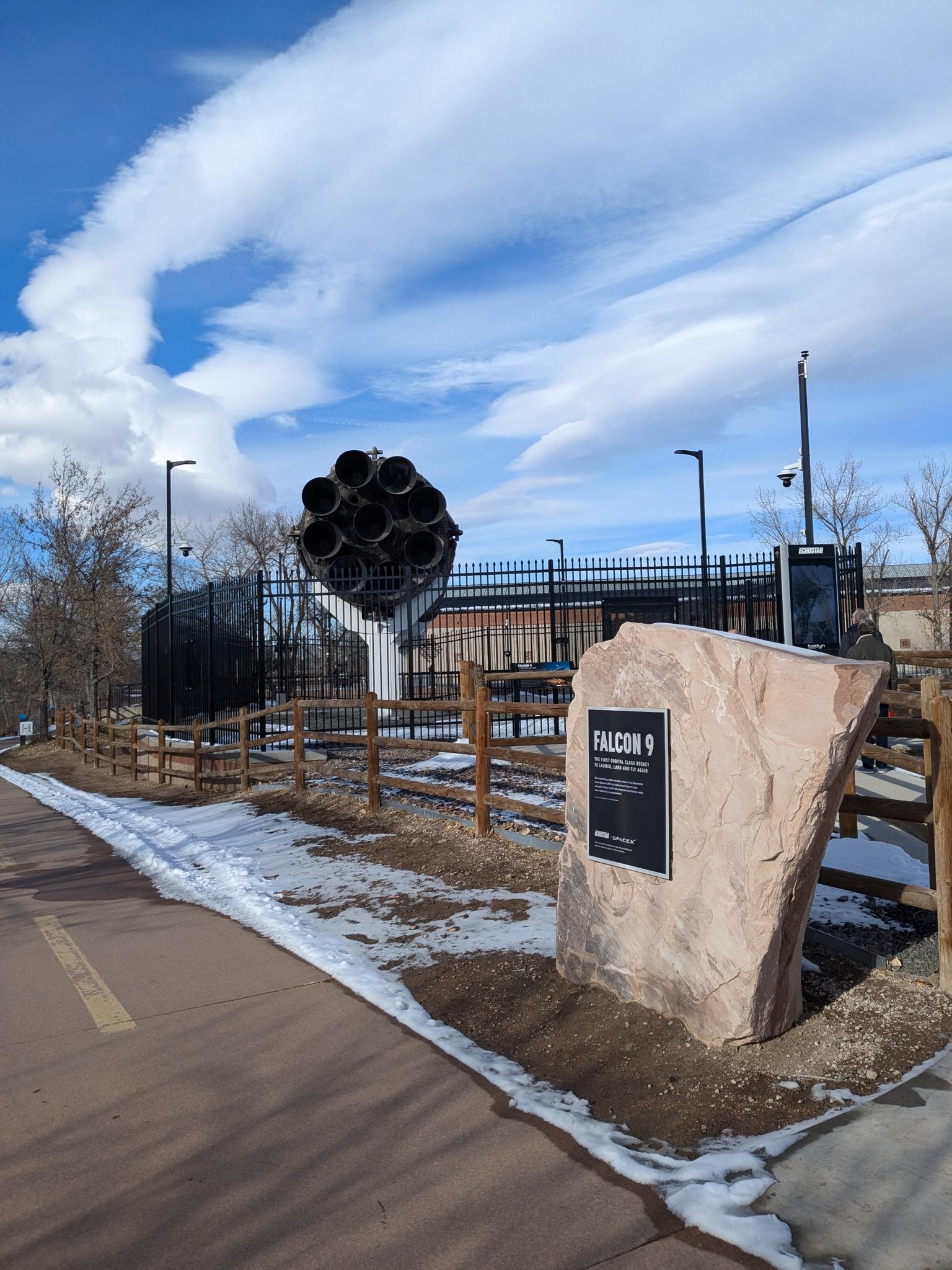 Large rocket engine display next to a paved path with snow patches.