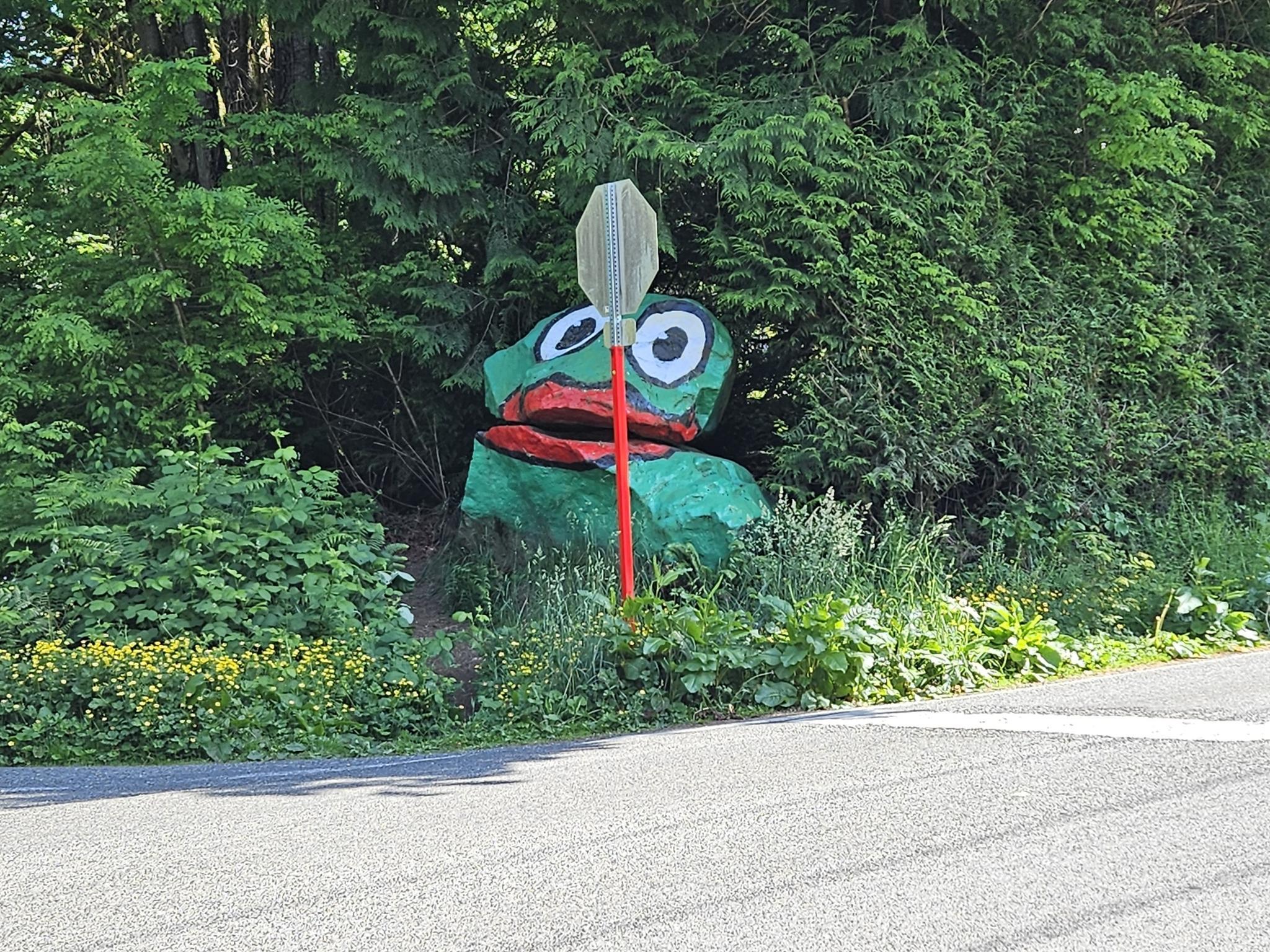 A large green frog sculpture with a stop sign by a road.