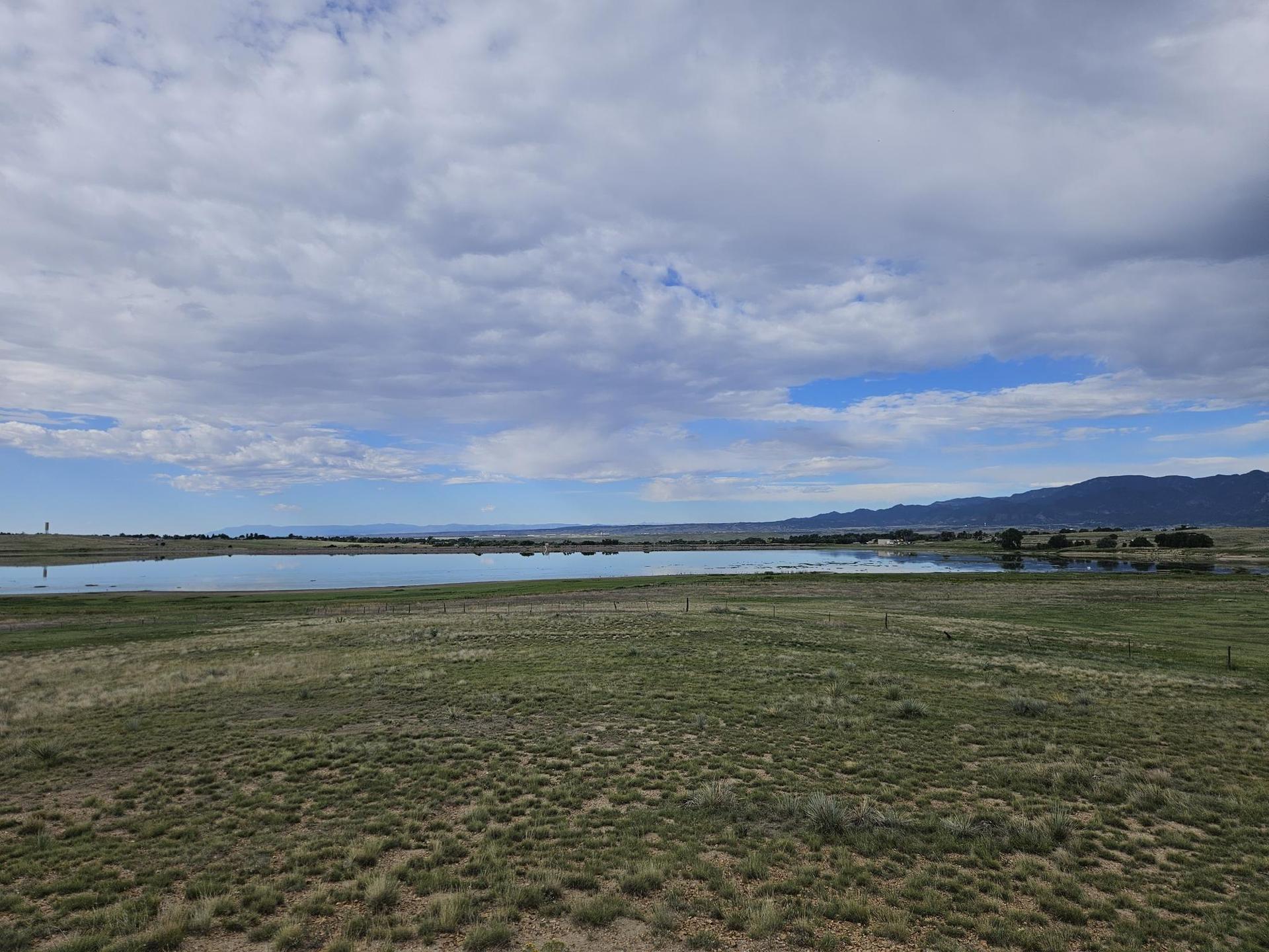 Calm lake reflecting a cloudy sky, surrounded by vast green fields and distant mountains.