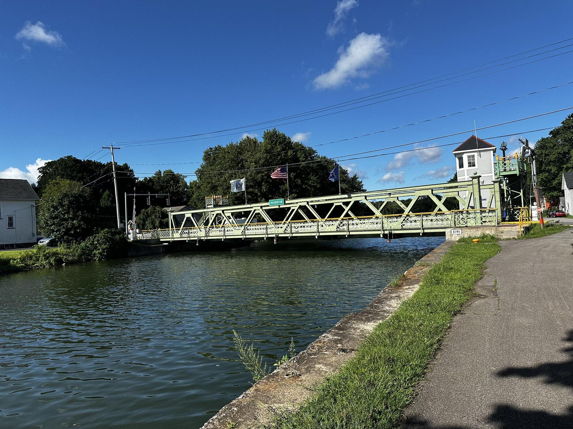 A green truss bridge spans a canal on a clear, sunny day.