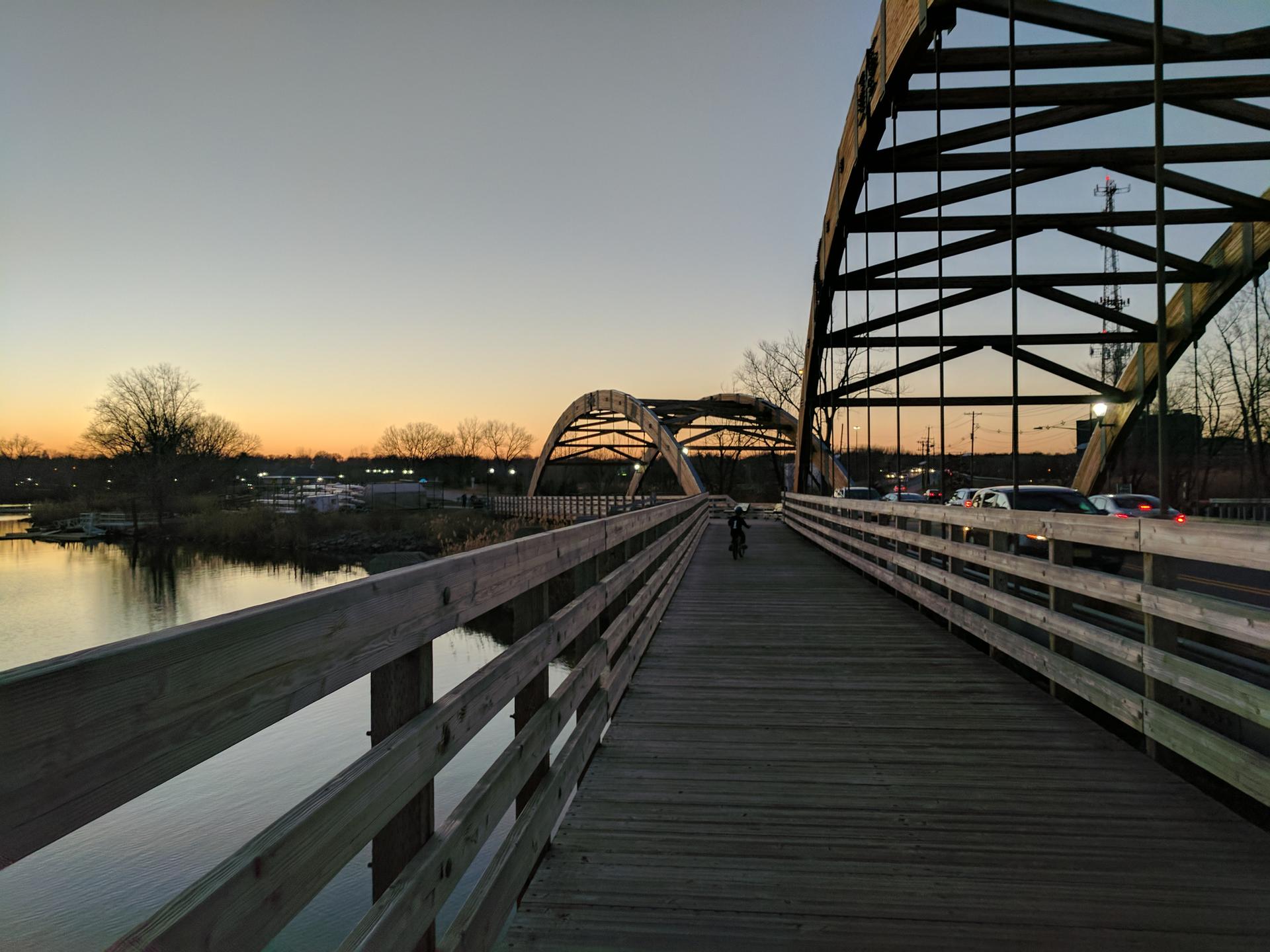 Wooden pedestrian bridge next to an arched metal bridge at sunset with a cyclist.