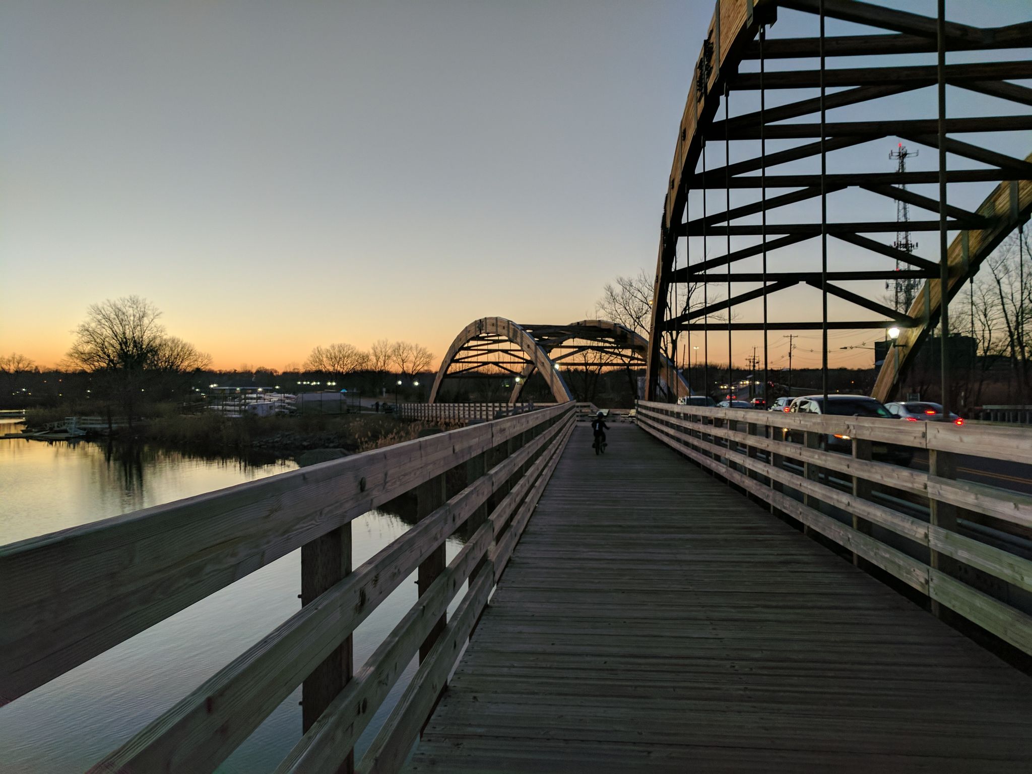 Wooden pedestrian bridge next to an arched metal bridge at sunset with a cyclist. - Overpeck Park loop