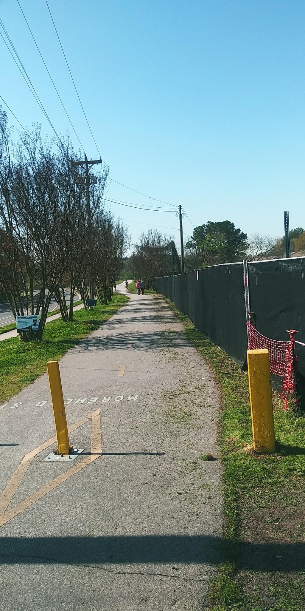 Paved path with yellow bollards, fence, and distant person under clear sky.