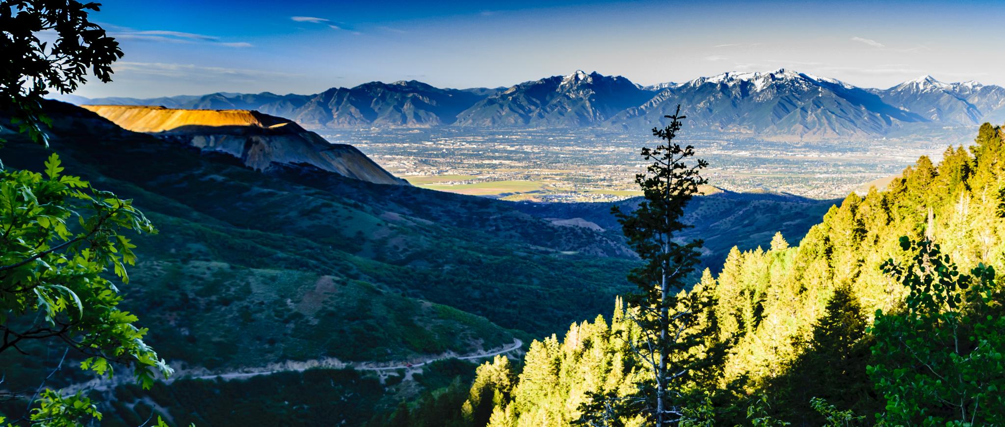 Panoramic view of a green valley, distant city, and snow-capped mountains under bright sun.