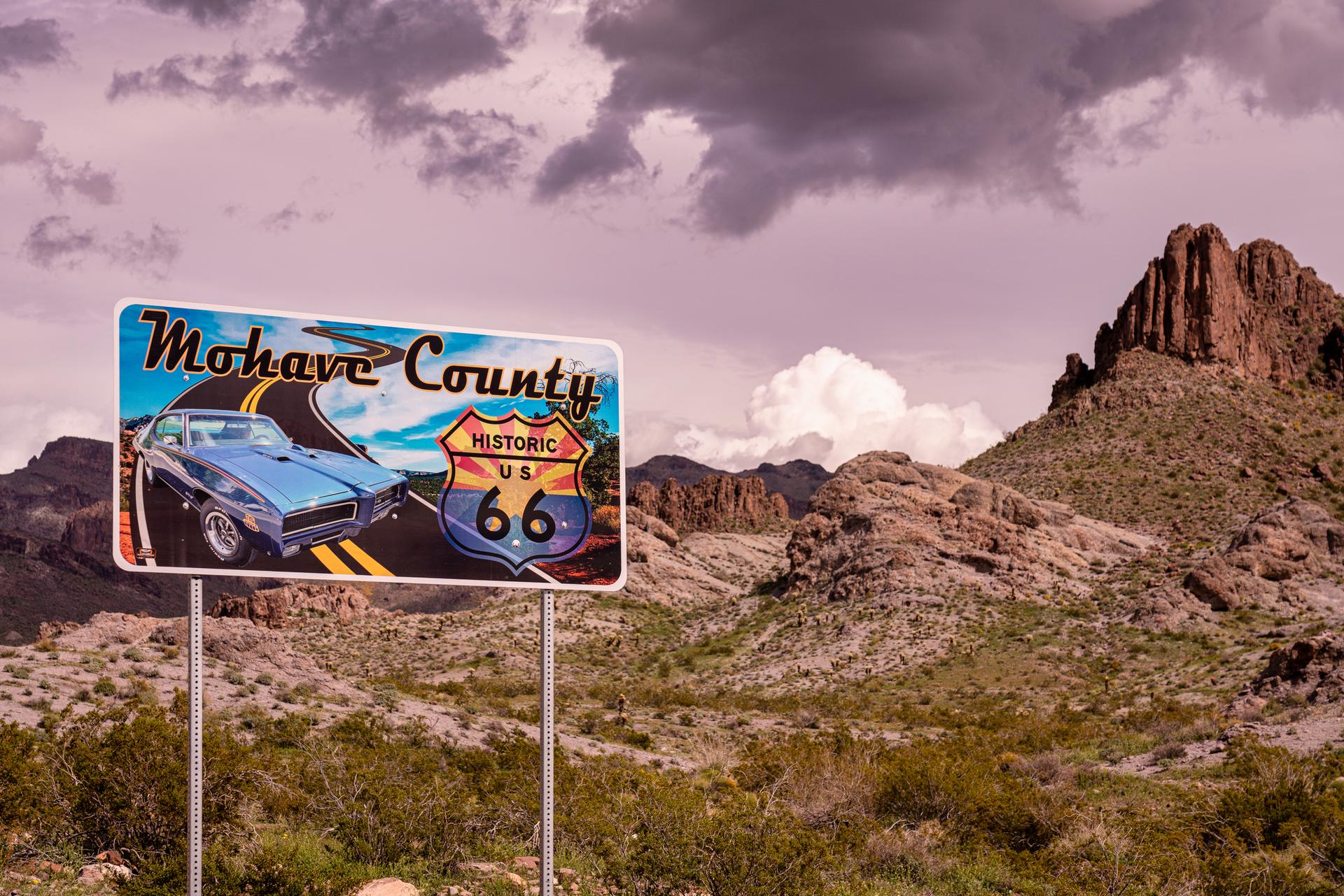 Road sign with a classic car and desert mountains under a cloudy sky.