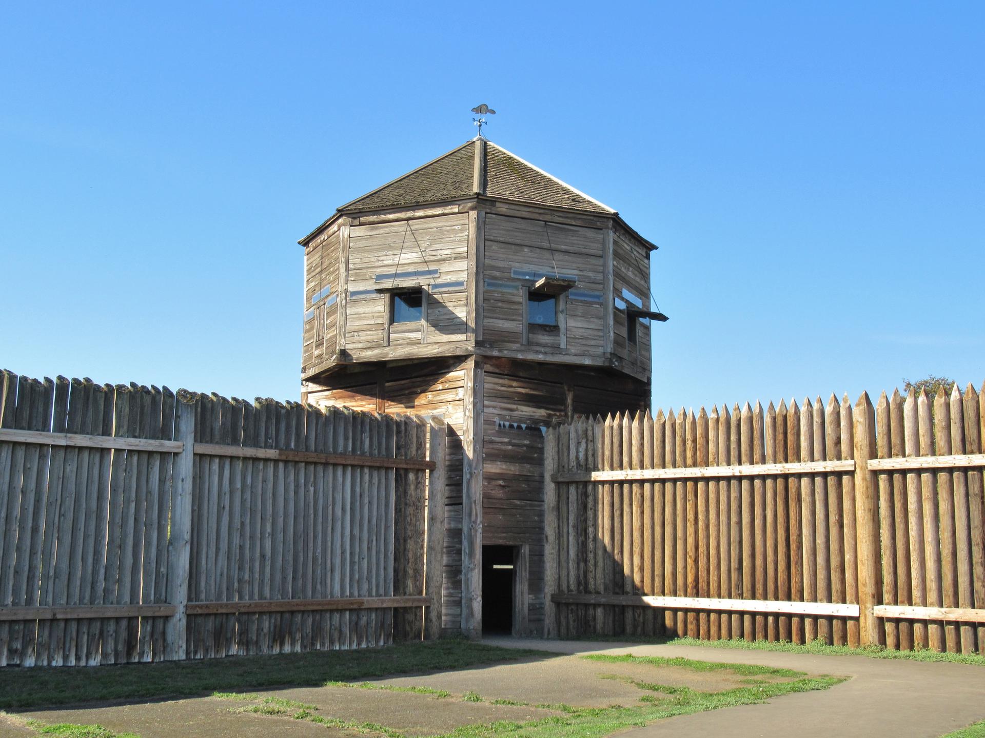 A wooden blockhouse stands within a tall wooden palisade fence.